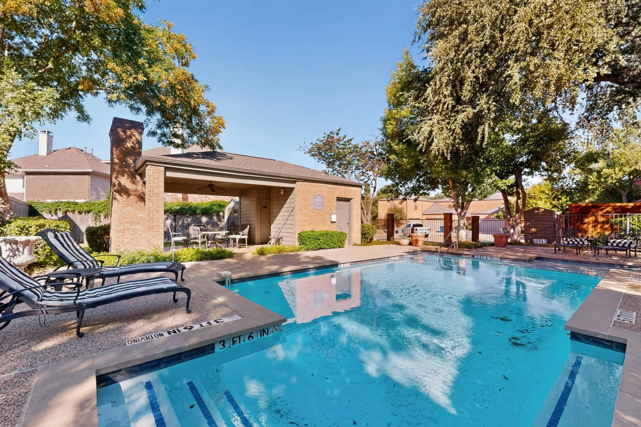 Community Resort-style pool area featuring a brick-clad covered patio, integrated pool steps, concrete pool deck, and mature trees