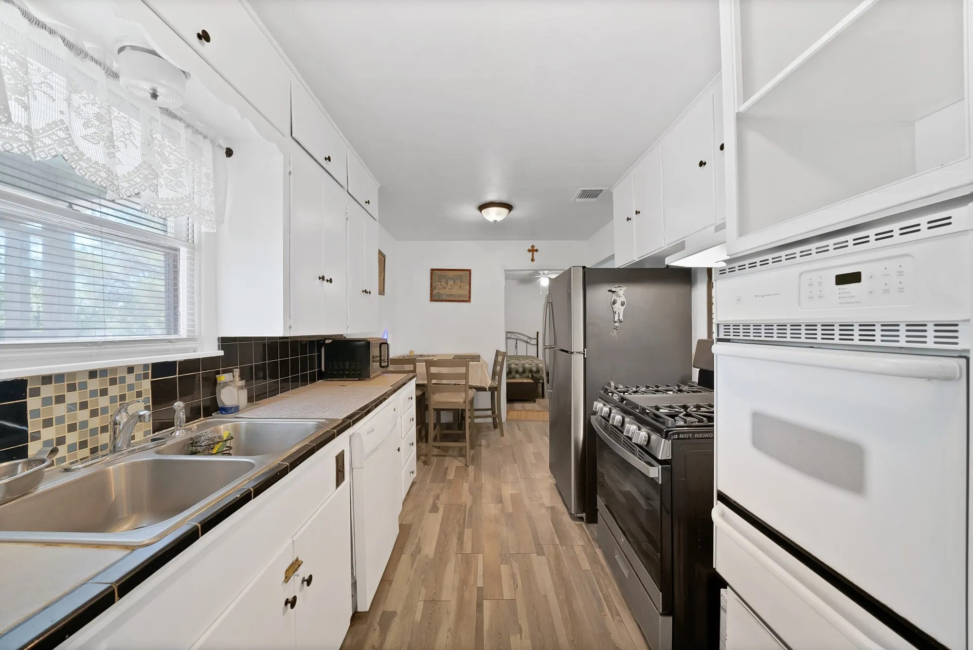 Galley kitchen featuring white cabinetry, wood-finish flooring, a stainless steel double sink, and a multi-tone tile backsplash