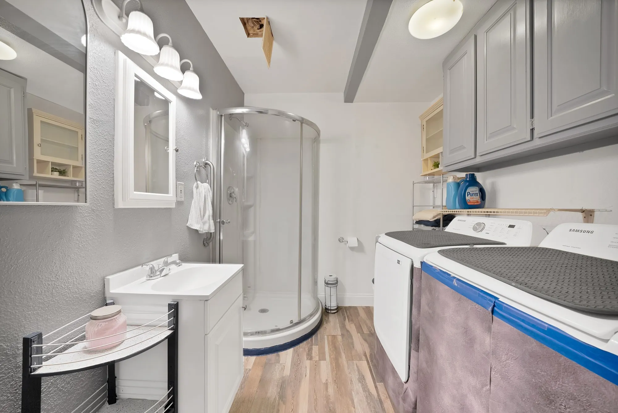 Vanity with single-basin sink, corner glass-enclosed shower, wood-finish flooring, and overhead cabinetry