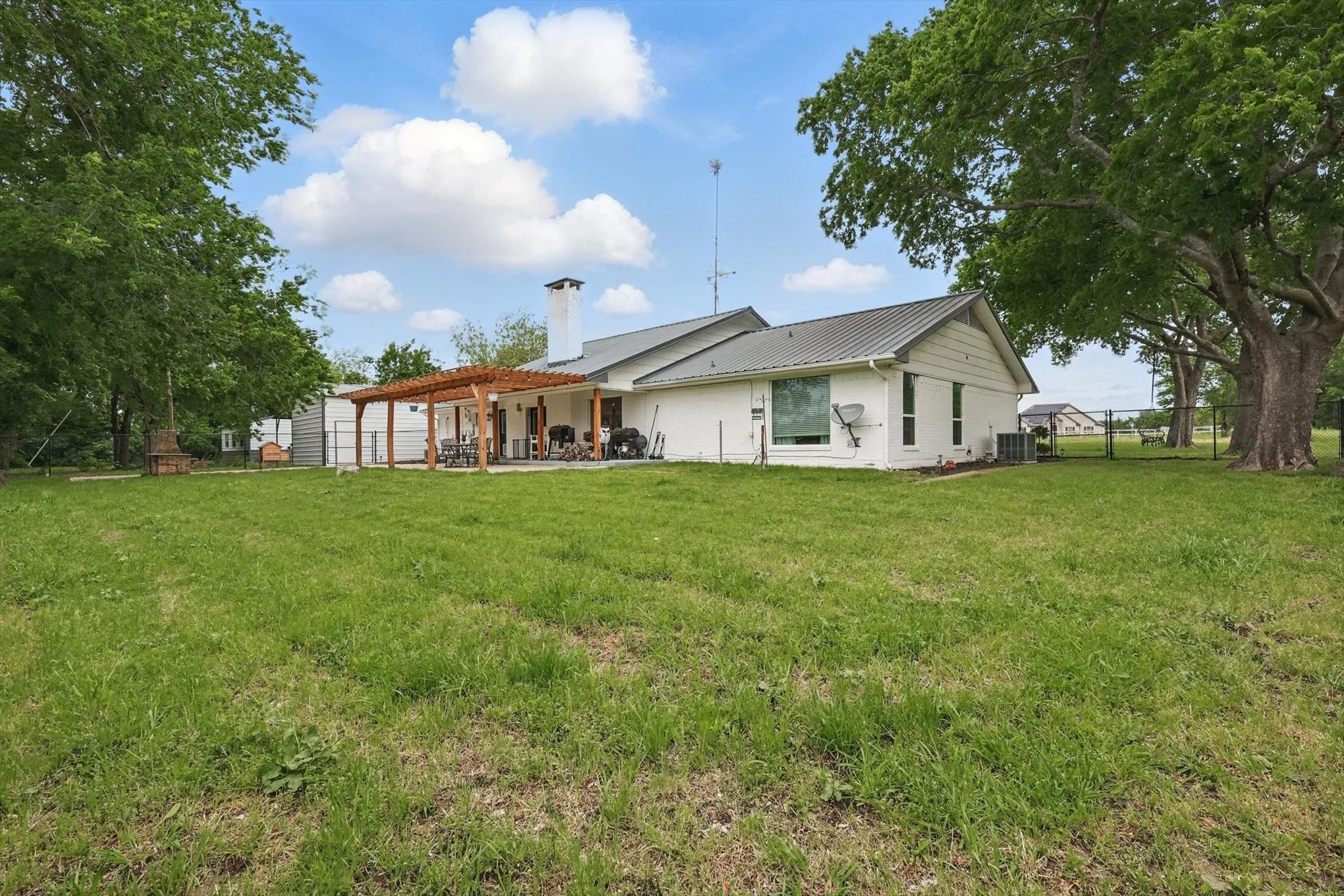 Expansive green space featuring a white exterior residence with a gray metal roof, a wood pergola over a concrete patio, and mature trees