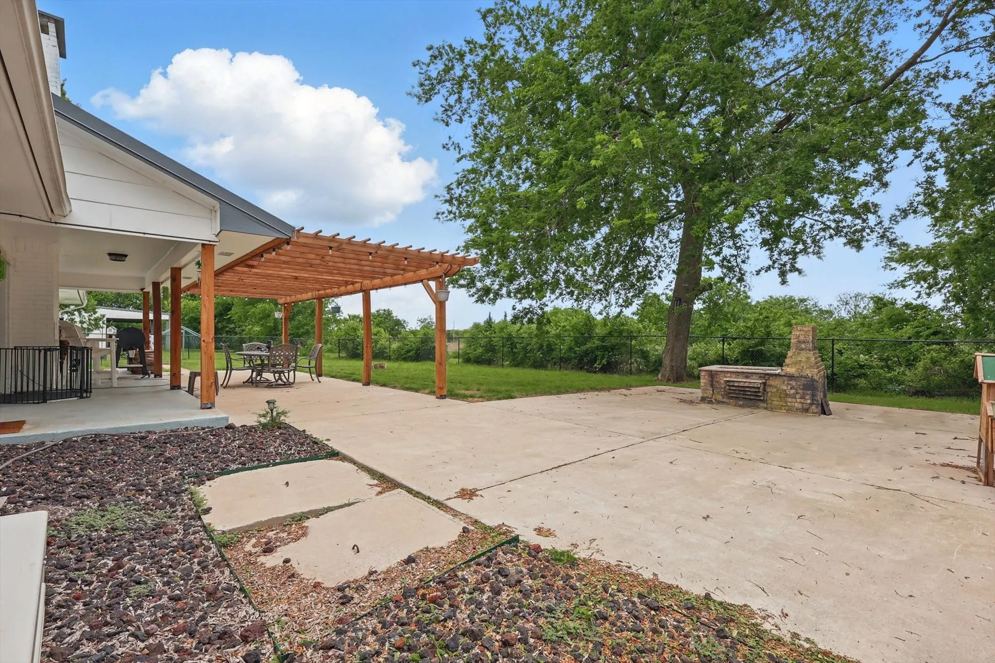 Expansive concrete patio with mature shade trees and open lawn space, complemented by a cedar pergola structure attached to the home