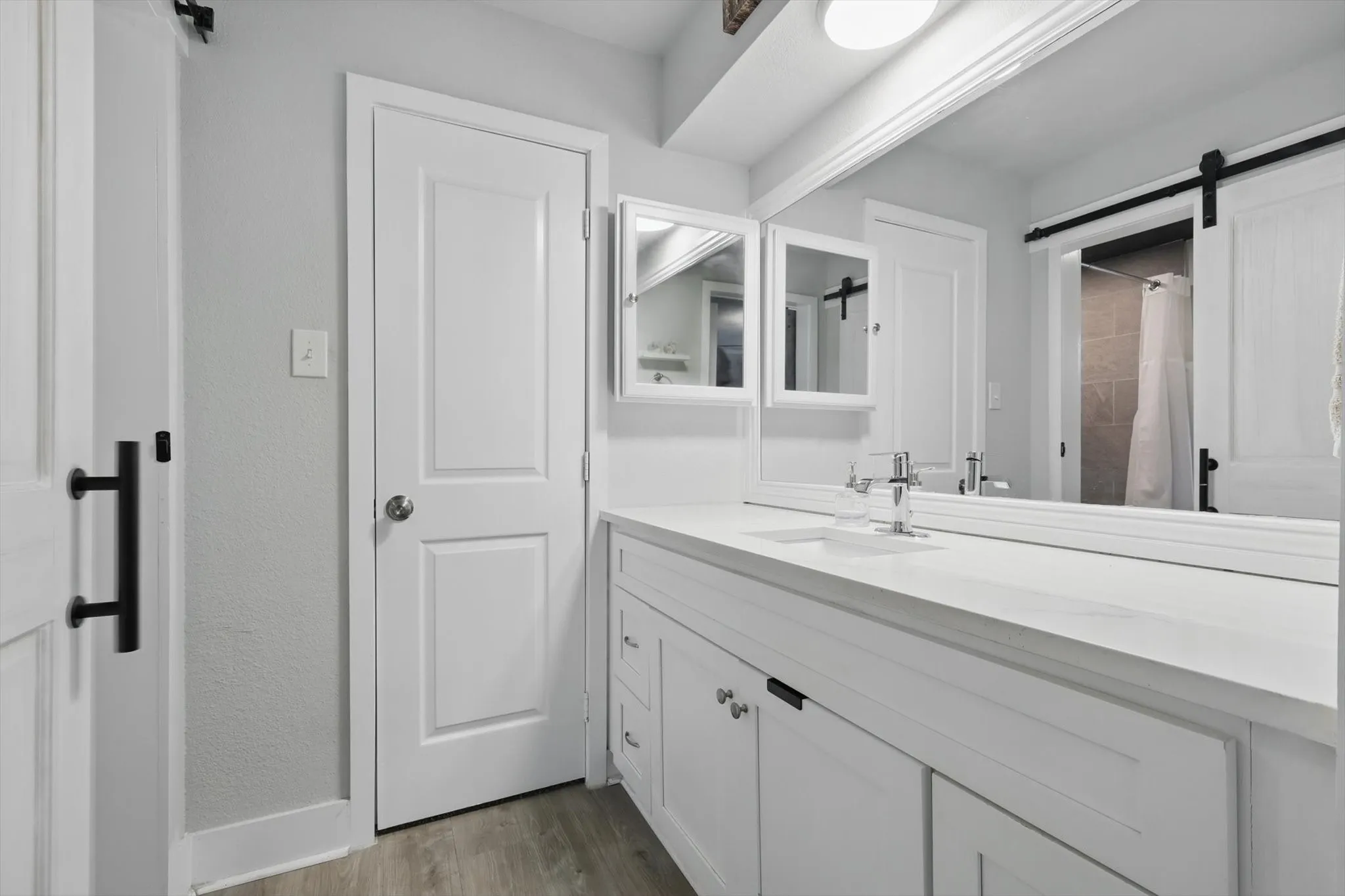 Modern primary bathroom featuring dual vanities with white cabinetry, an extended countertop with integrated sinks, chrome fixtures, large framed mirror, recessed and linear ceiling lighting, wood-finish flooring, and direct access to the shower area