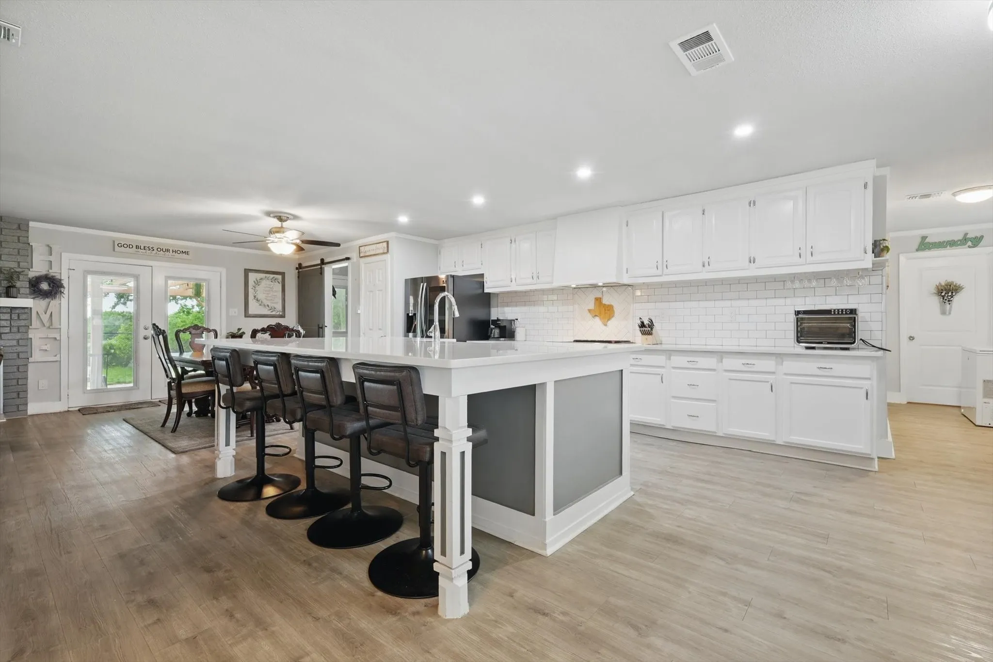 Open-concept kitchen and dining area featuring white cabinetry with brushed nickel hardware, a white kitchen island with gray lower panels and seating for multiple guests, wood-finish flooring throughout, stainless steel appliances, subway tile backsplash, and recessed ceiling lighting