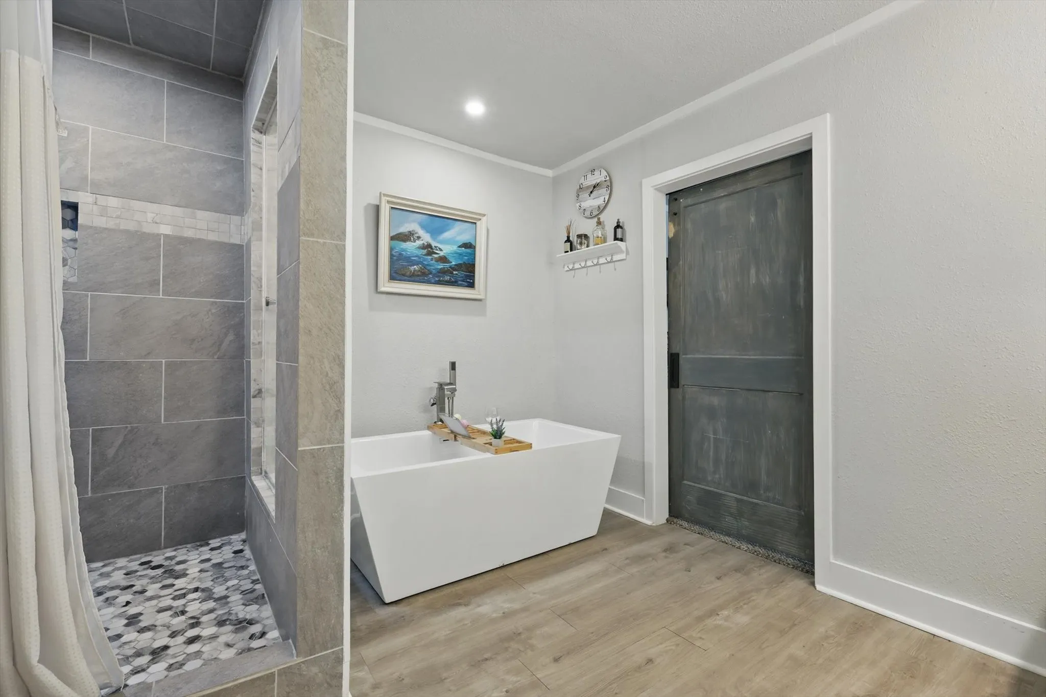 Spa-inspired primary bathroom featuring a freestanding soaking tub, walk-in shower with large-format tile surround and mosaic floor detail, wood-finish flooring, recessed lighting, and soft gray wall finishes complemented by coastal artwork