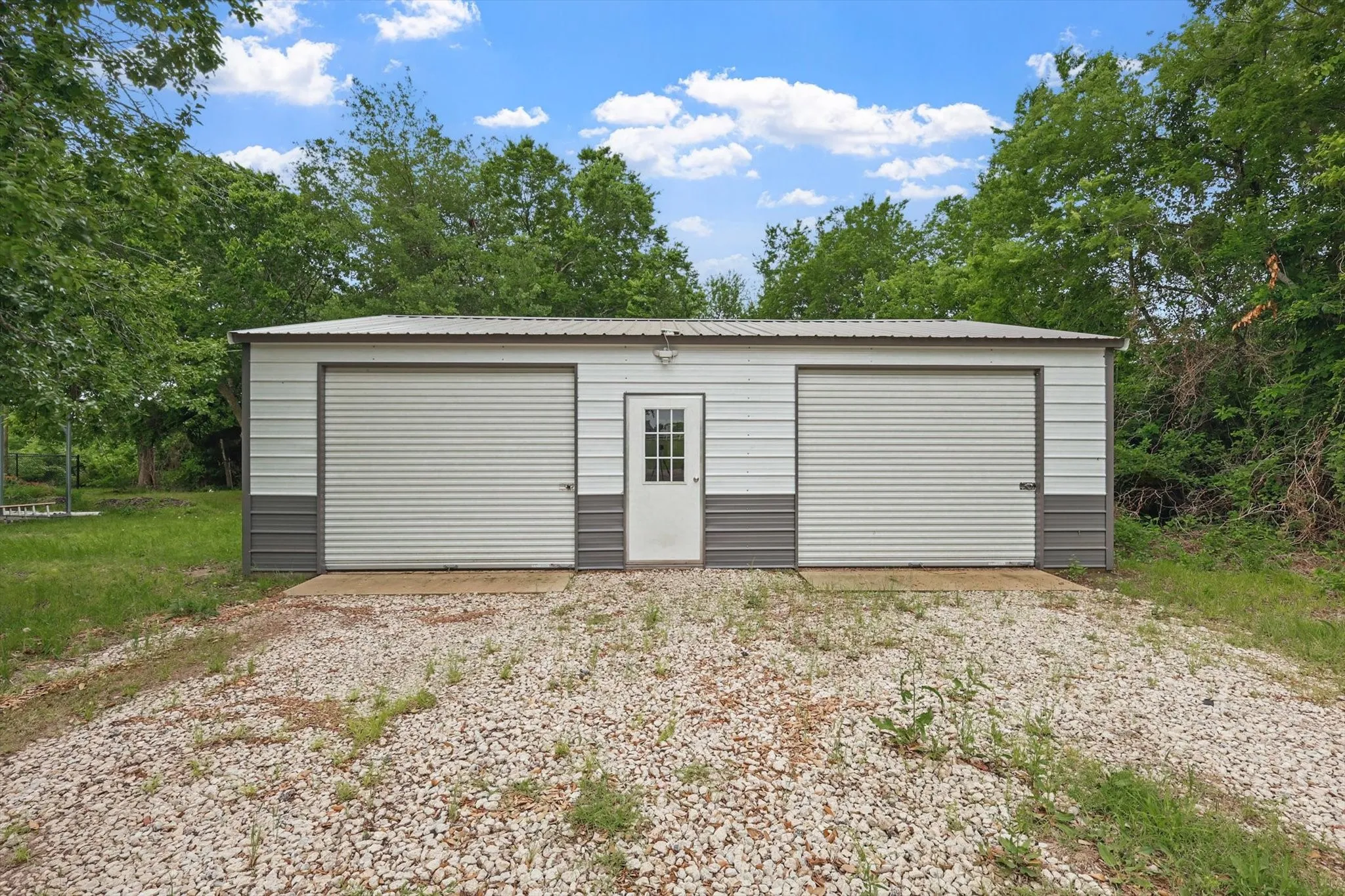 Metal storage building with two roll-up garage doors, a pedestrian entry door, and a gravel driveway