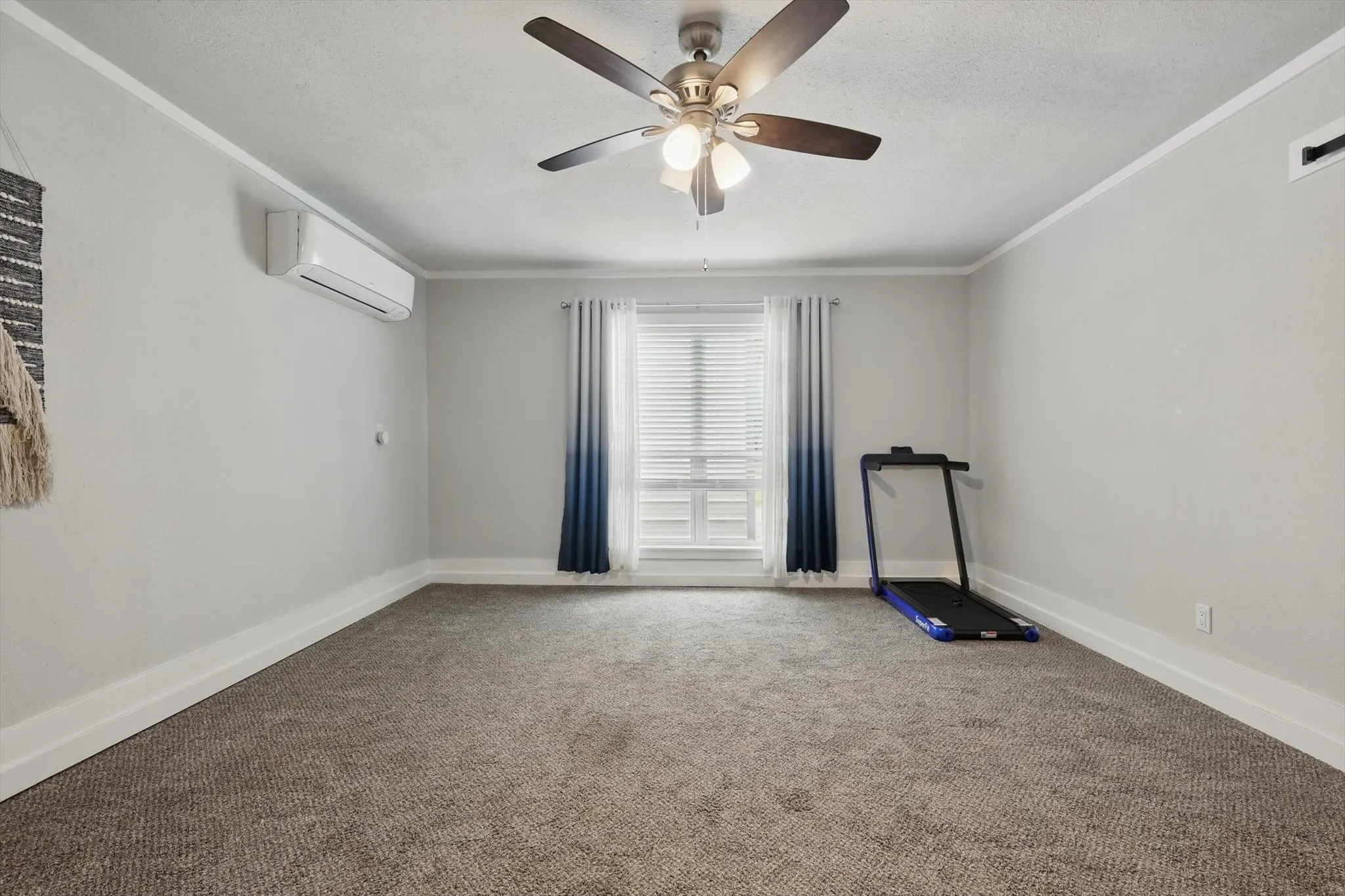 Neutral wall paint, ceiling fan with integrated lighting, window with blinds, wall-mounted air conditioning unit, and carpet flooring