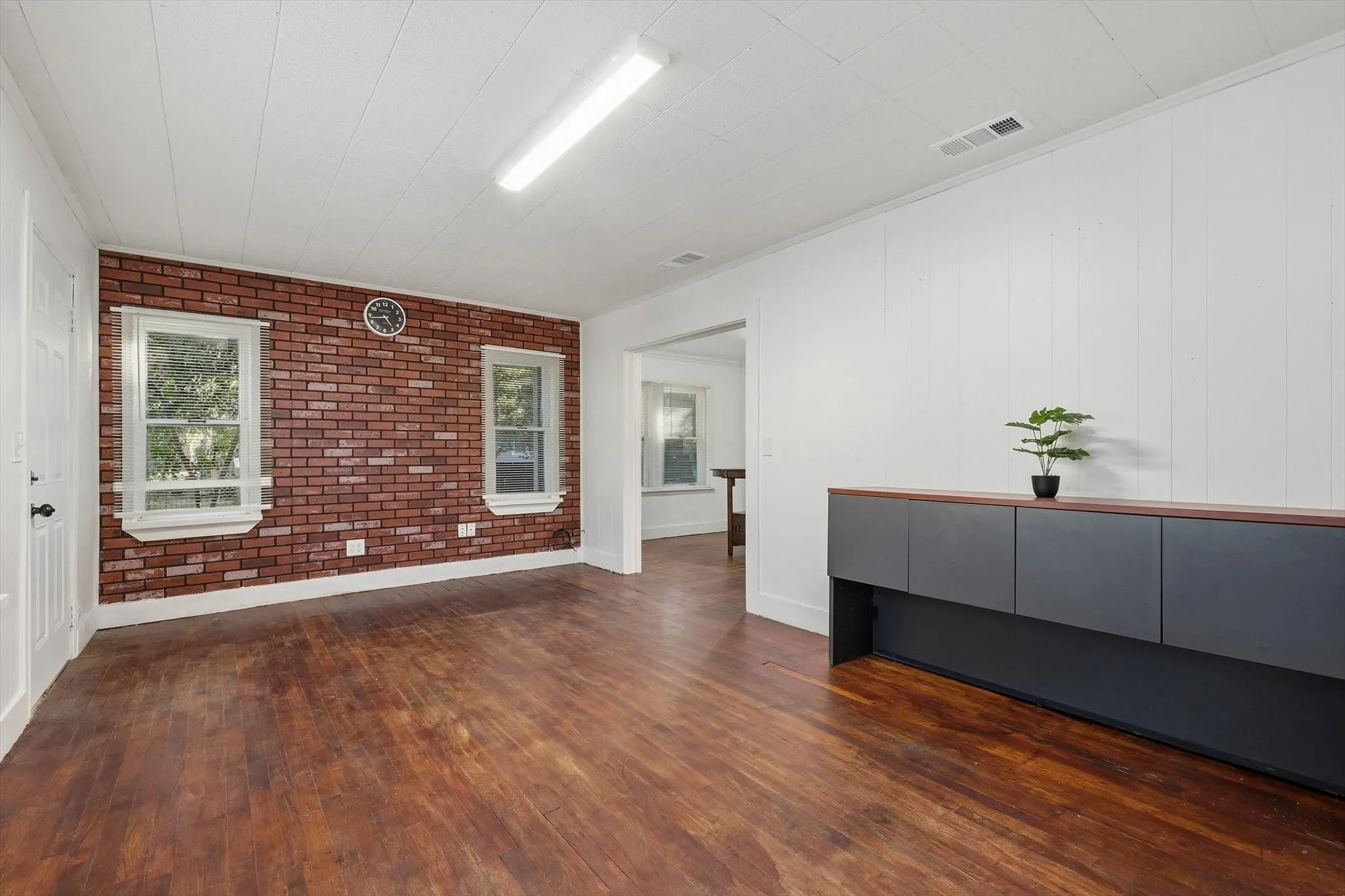 Wood-finish flooring, accent brick wall, white paneled walls, white ceiling, and a built-in dark cabinetry with a wood-finish top