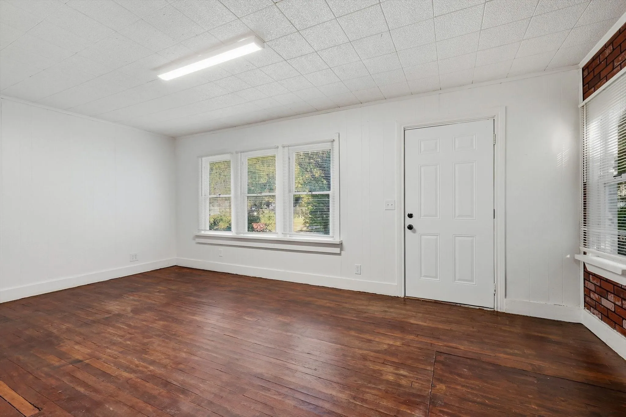 Wood-finish flooring, white paneled walls, a white paneled door, three large windows, and a brick accent wall