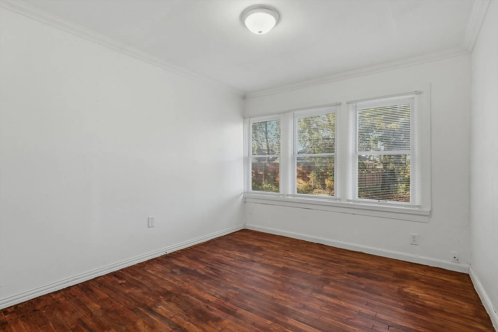 Wood-finish flooring, white walls, triple window configuration, white window blinds, and a flush-mount ceiling light