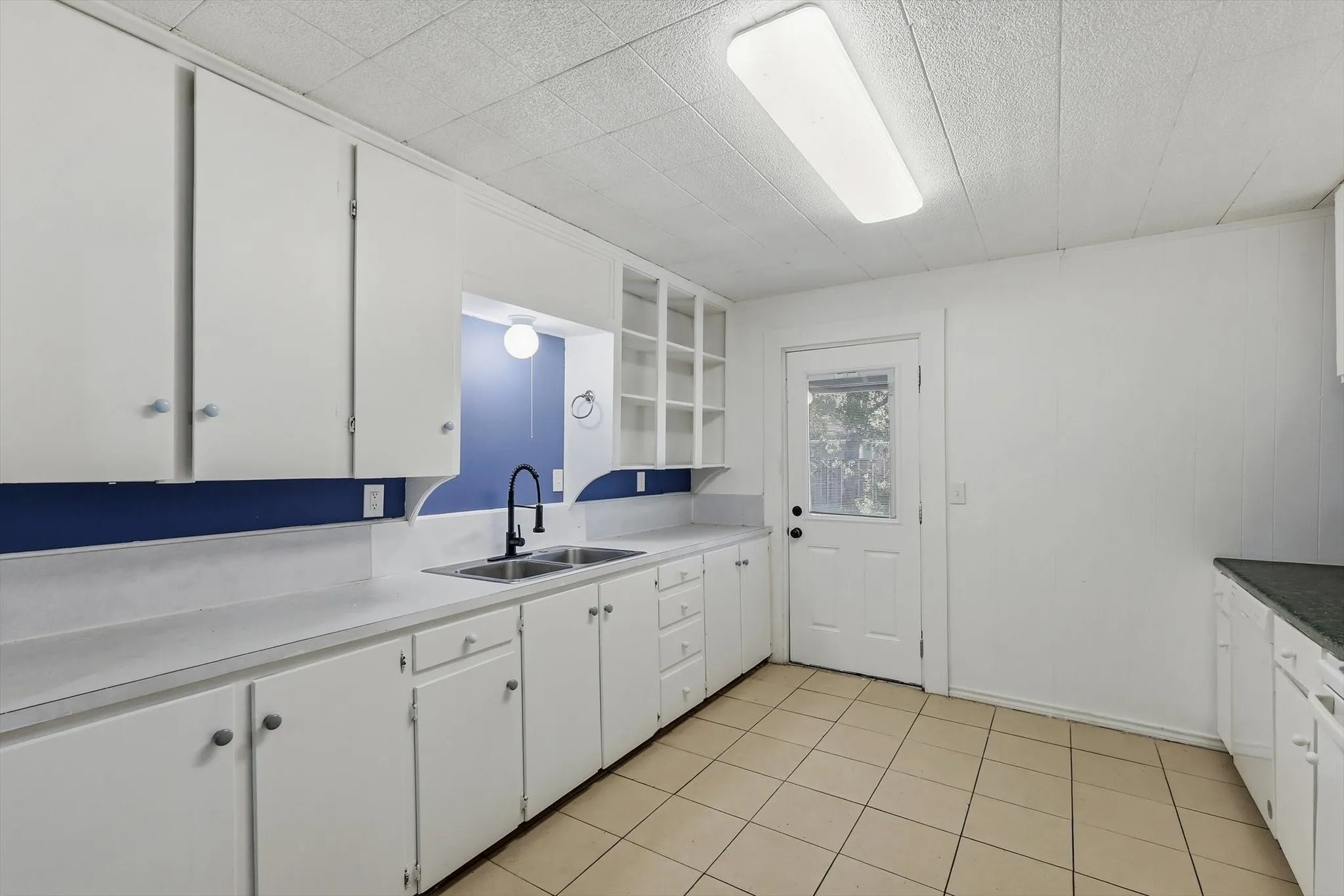 White cabinetry, dual basin stainless steel sink, black gooseneck faucet, tiled flooring, and a white back door with a window