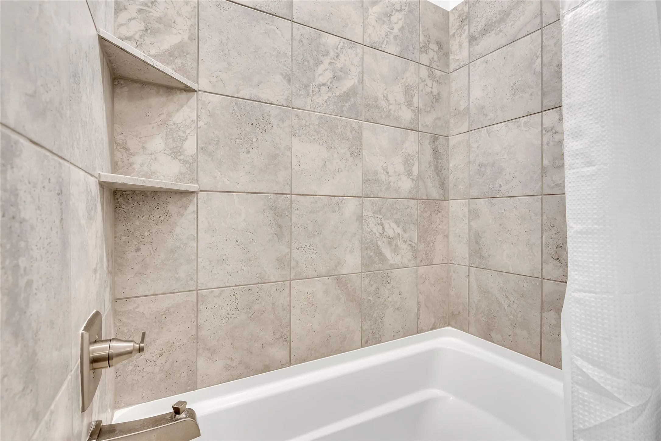 Bathtub with neutral-toned wall tiles, two recessed corner shelves, and brushed metal fixtures