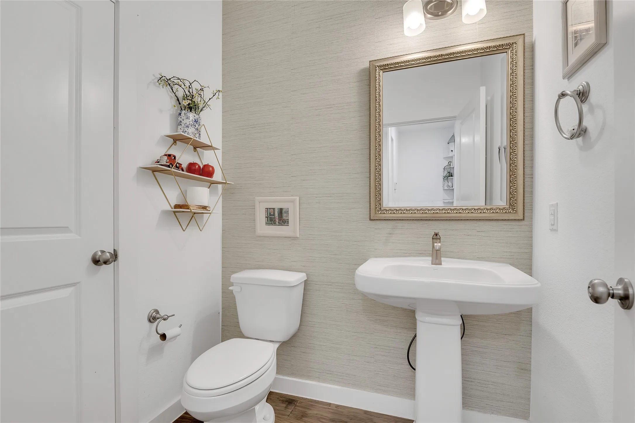 Pedestal sink, brushed nickel faucet, framed mirror, wall-mounted light fixture, and wood-finish flooring