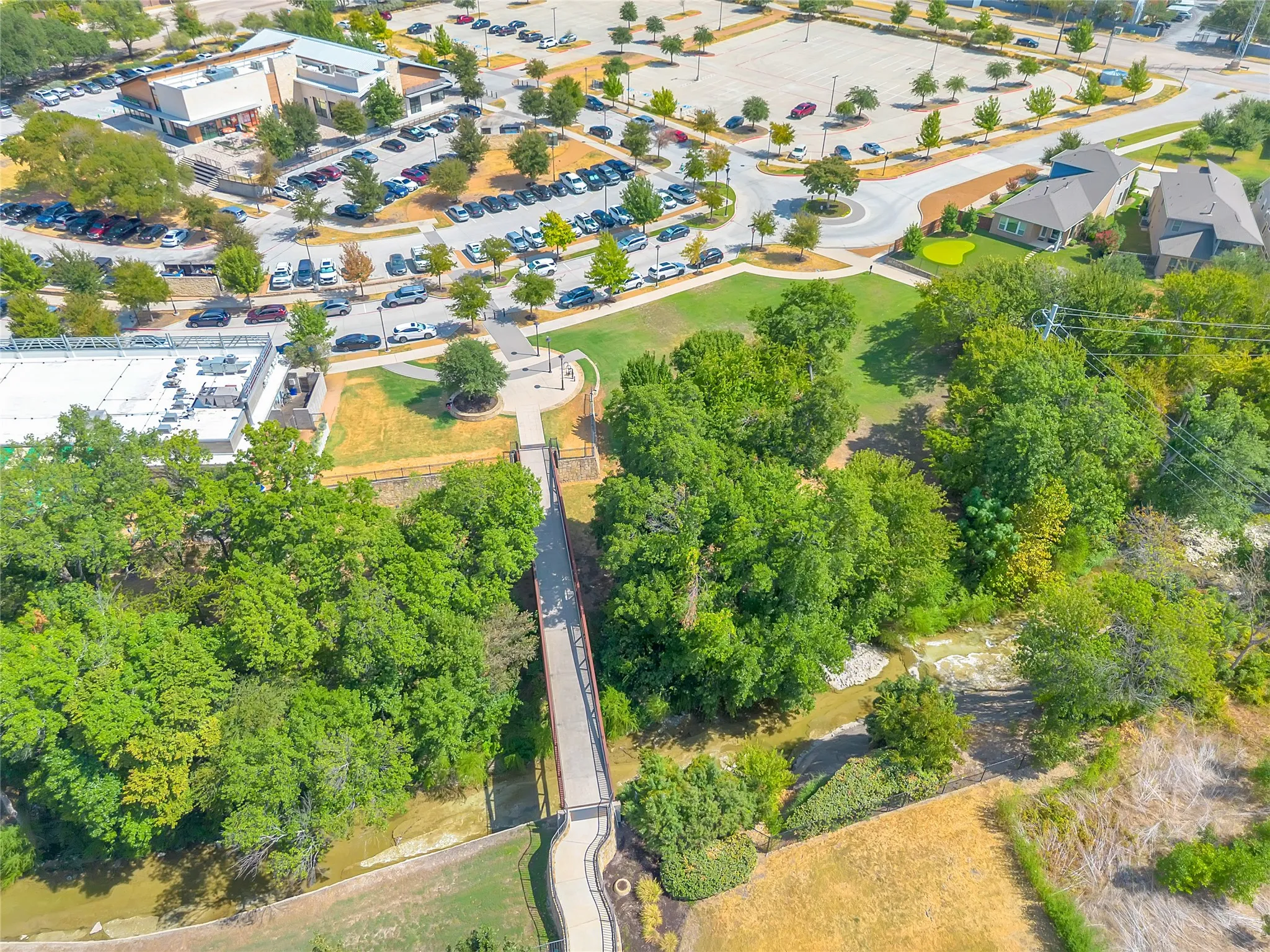 Aerial perspective highlighting a pedestrian bridge spanning a natural waterway, surrounded by mature trees and manicured green spaces