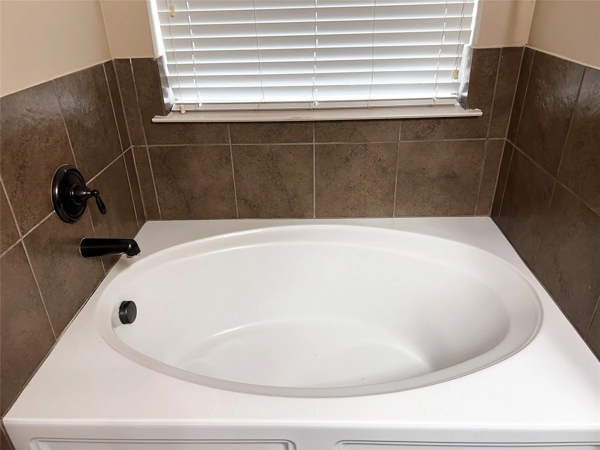 Oval soaking tub with a tiled surround, oil-rubbed bronze fixtures, and white window blinds