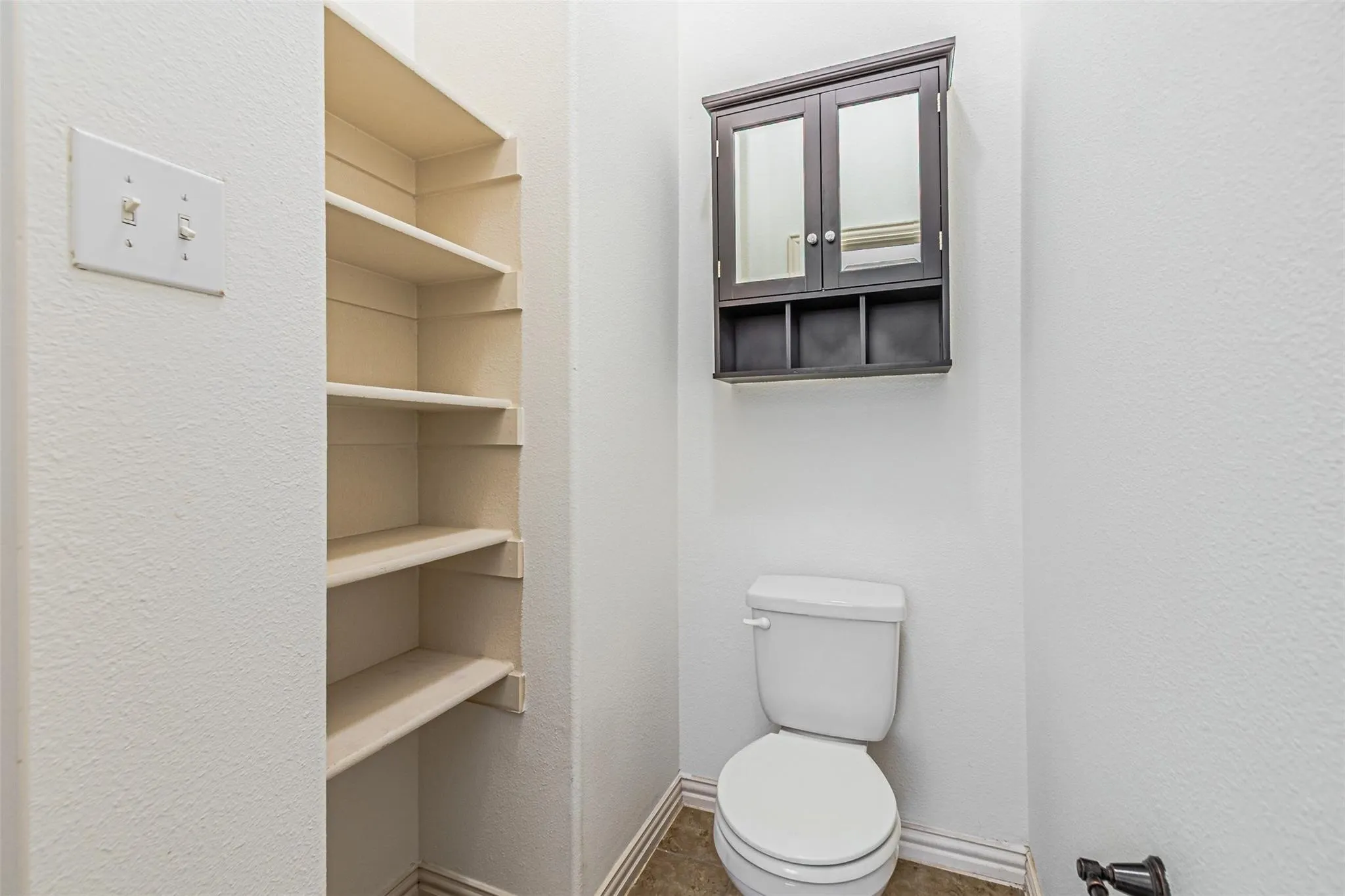 Built-in shelving, dark wood-finish medicine cabinet with mirrored doors, and a toilet with a white finish