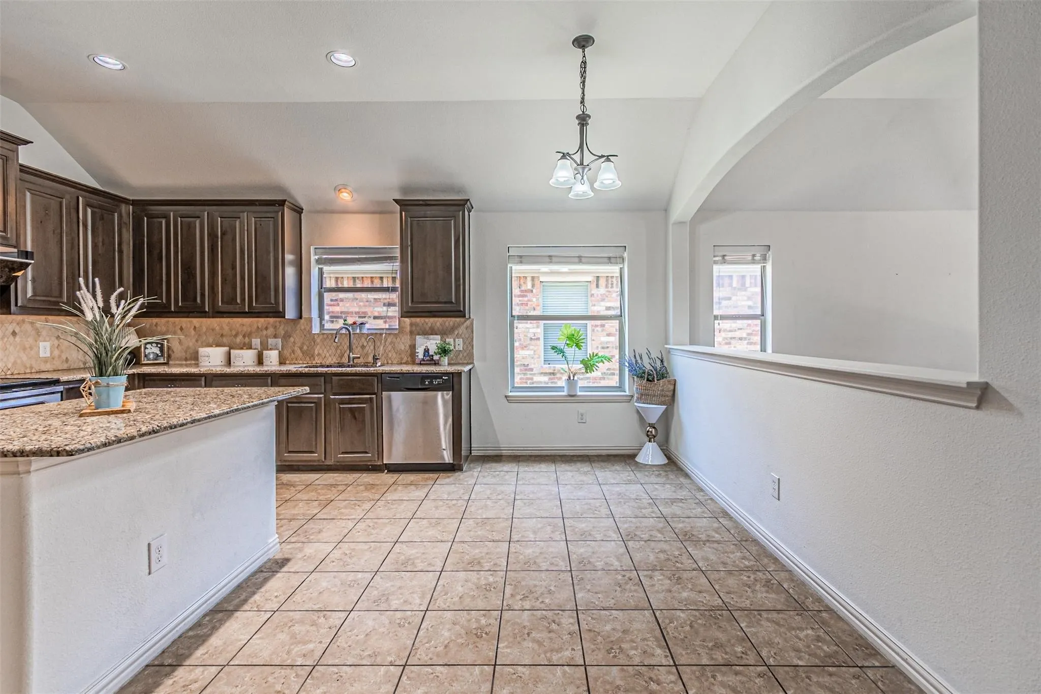 Spacious kitchen and dining area featuring wood-finish cabinetry with decorative detailing, granite countertops, stainless steel appliances, tile flooring throughout, recessed lighting, and pendant fixture