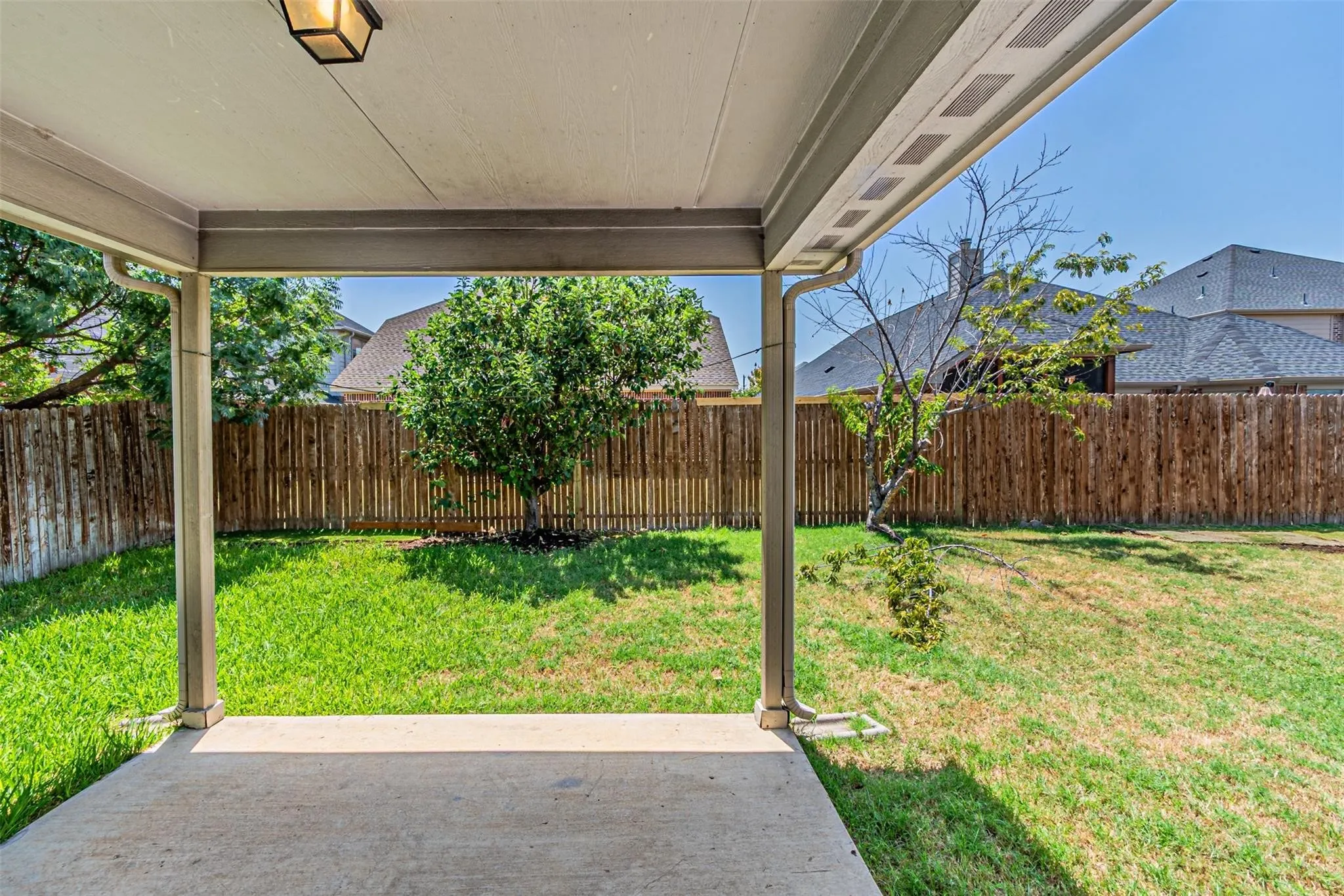 Covered patio with an outdoor light fixture, concrete flooring, and a fully fenced yard with mature trees