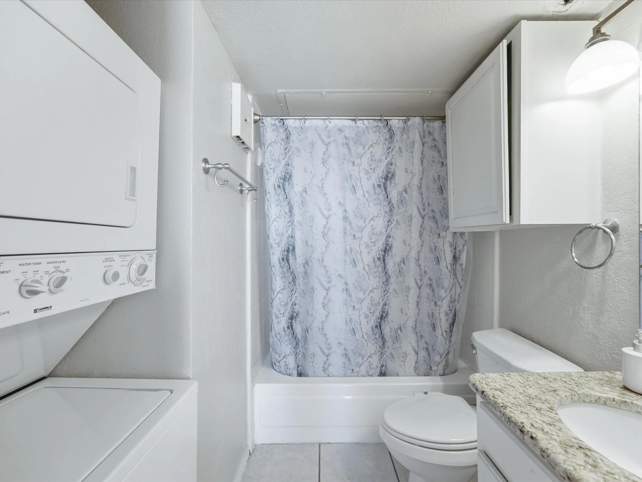 Bathroom featuring a white stacked washer and dryer, a tub-shower combination with a patterned curtain, a toilet, and a vanity with a light-colored granite countertop and integrated sink