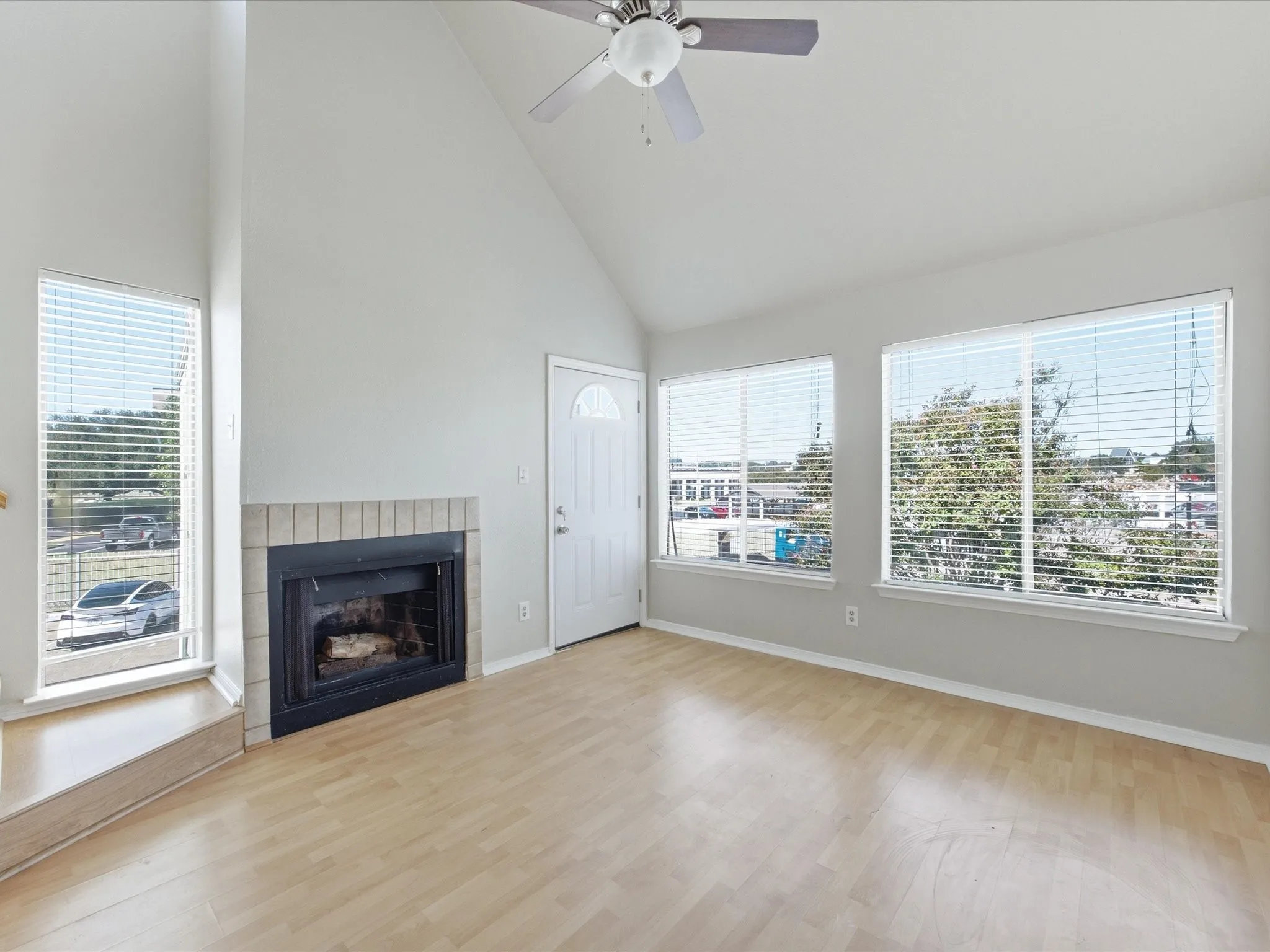 Vaulted ceiling, wood-finish flooring, fireplace with brick surround, multiple windows with blinds, and a ceiling fan