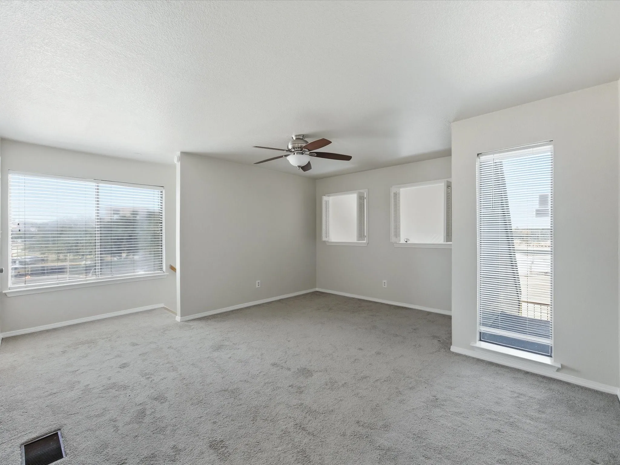 Spacious room featuring light gray carpeting, multiple windows with blinds, a ceiling fan, and white trim