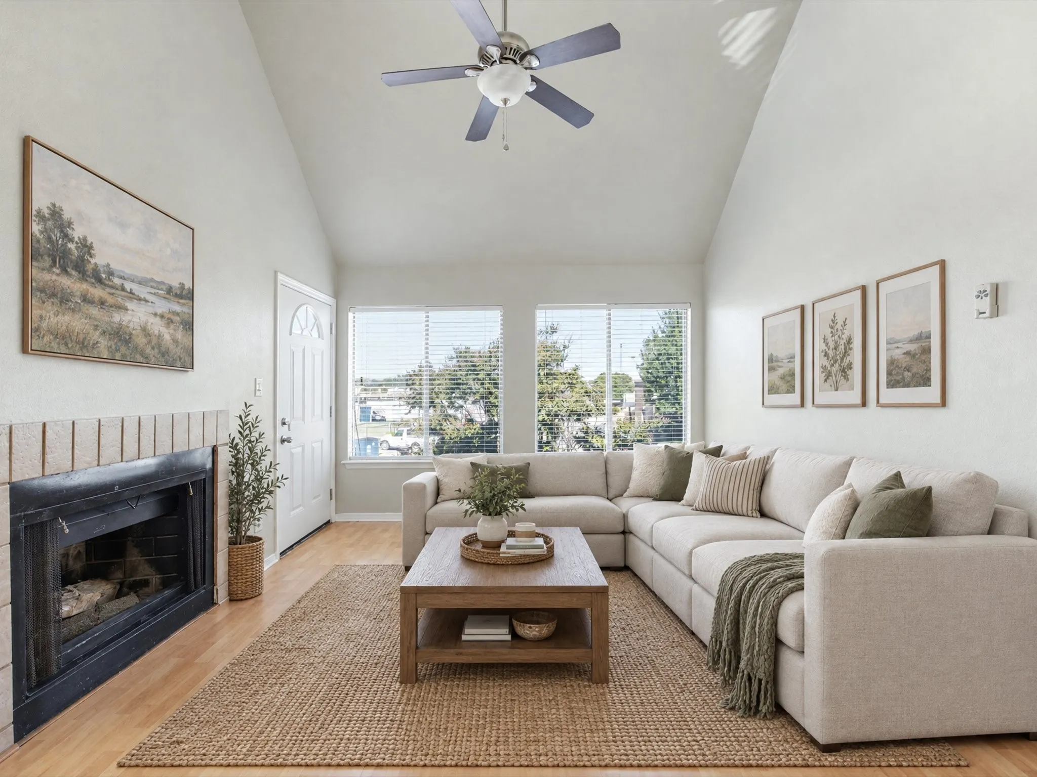 Vaulted ceiling with ceiling fan, wood-finish flooring, fireplace with brick surround, multiple windows, and a panel door with an arched glass insert