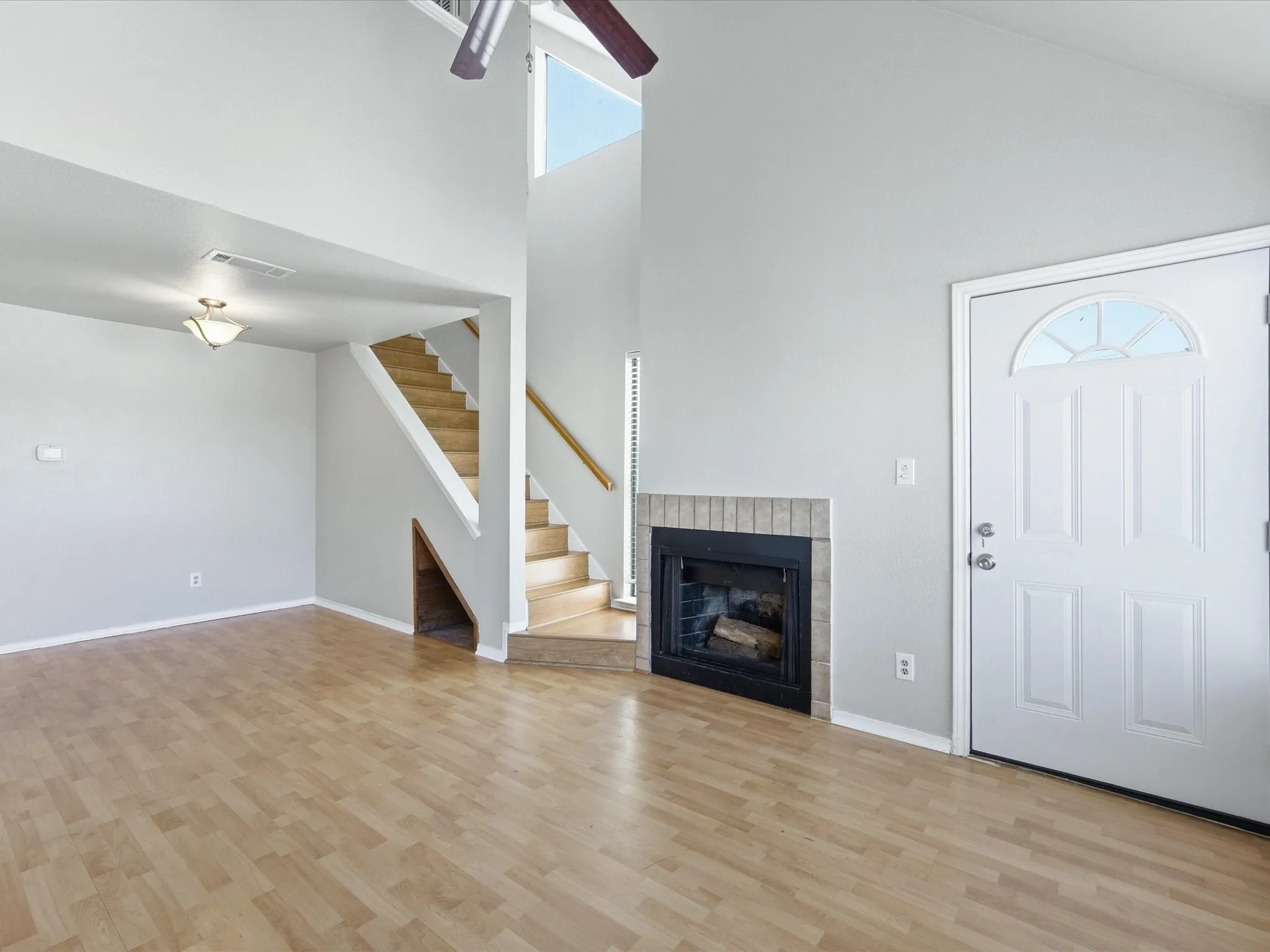 Vaulted ceilings with a ceiling fan, a fireplace with a brick surround, wood-finish flooring, a white paneled entry door with an arched window, and an open staircase with wood handrails
