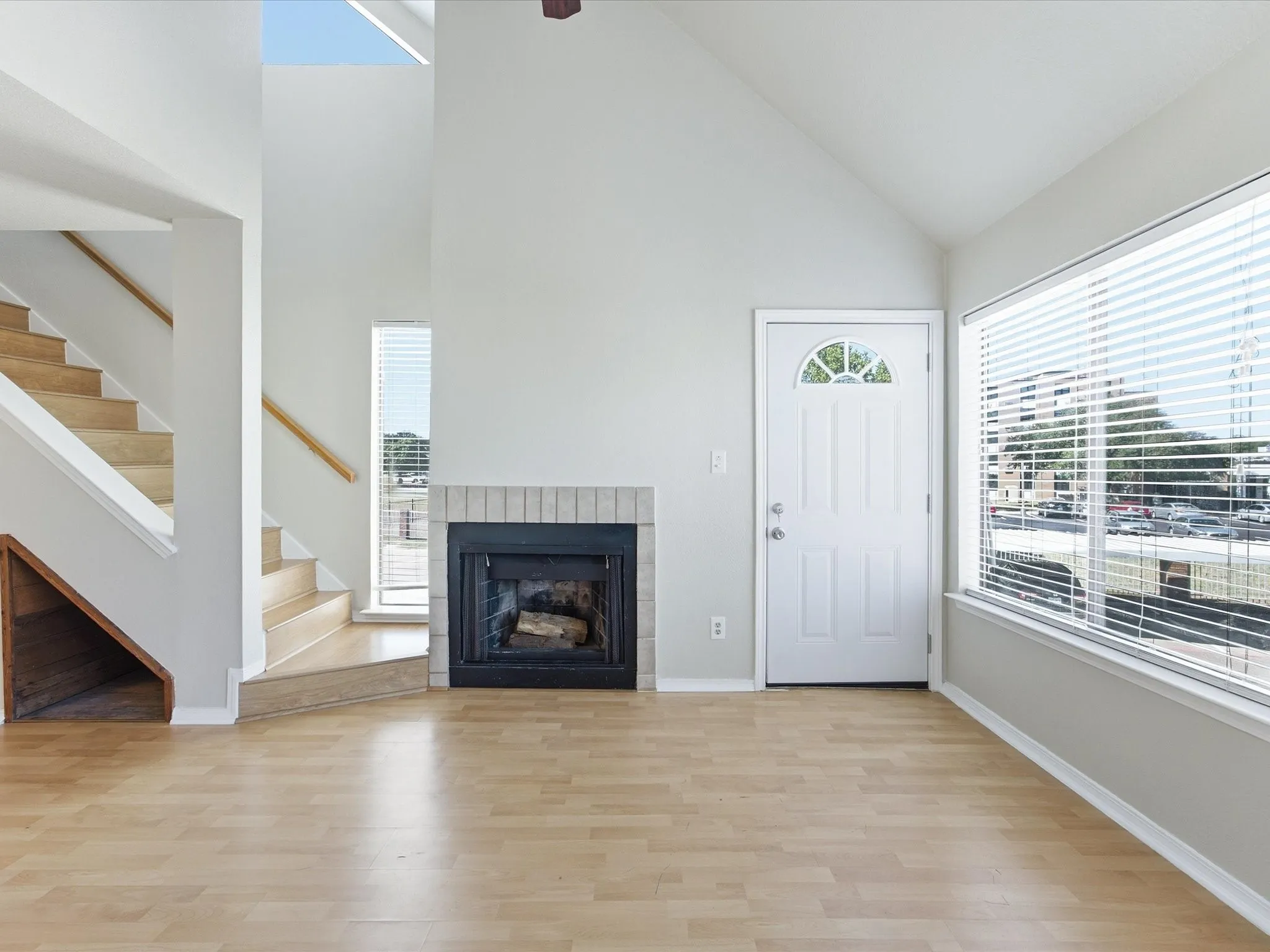 Vaulted ceiling, wood-finish flooring, brick-surround fireplace, large window with blinds, and a staircase with wood handrails