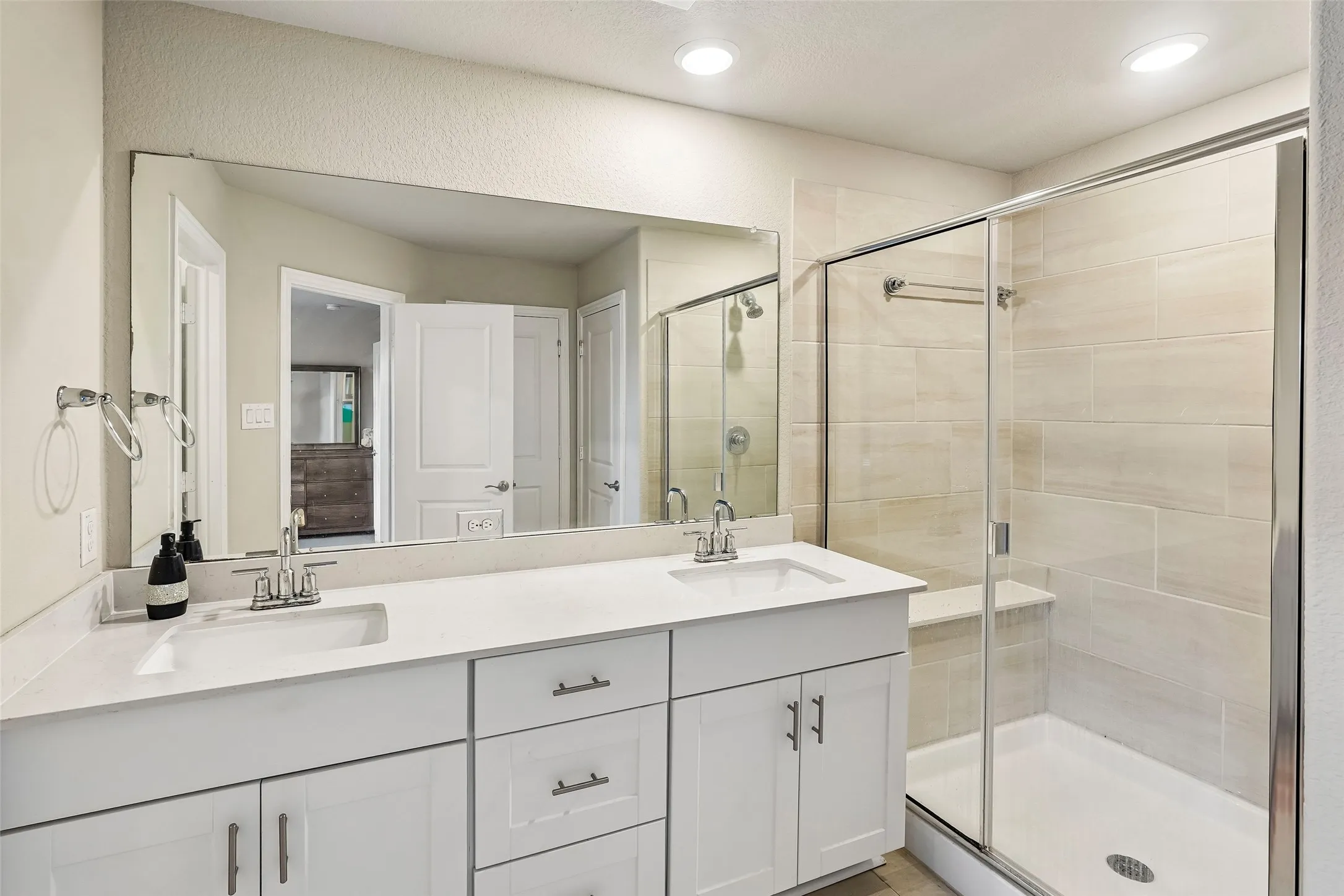 Dual vanity with integrated sinks, quartz countertop, white cabinetry with silver hardware, large frameless mirror, and a glass-enclosed shower with tiled walls