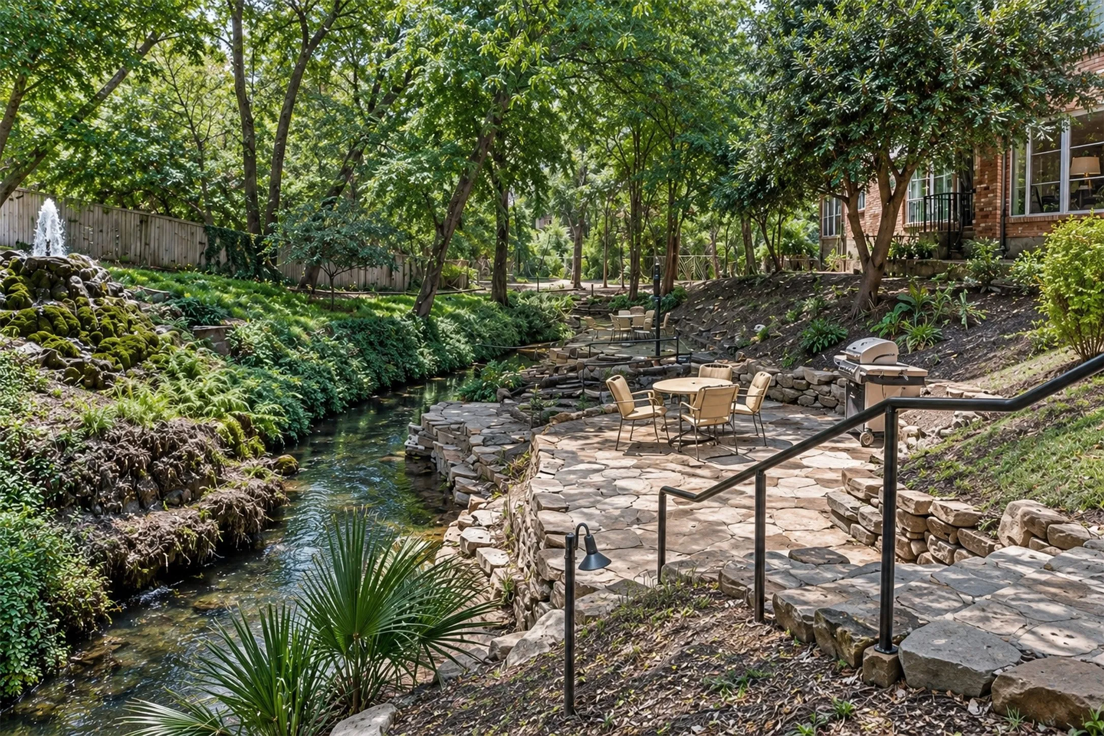 Expansive outdoor space featuring a natural stream, a decorative fountain, a stone patio with a built-in grill, and mature trees