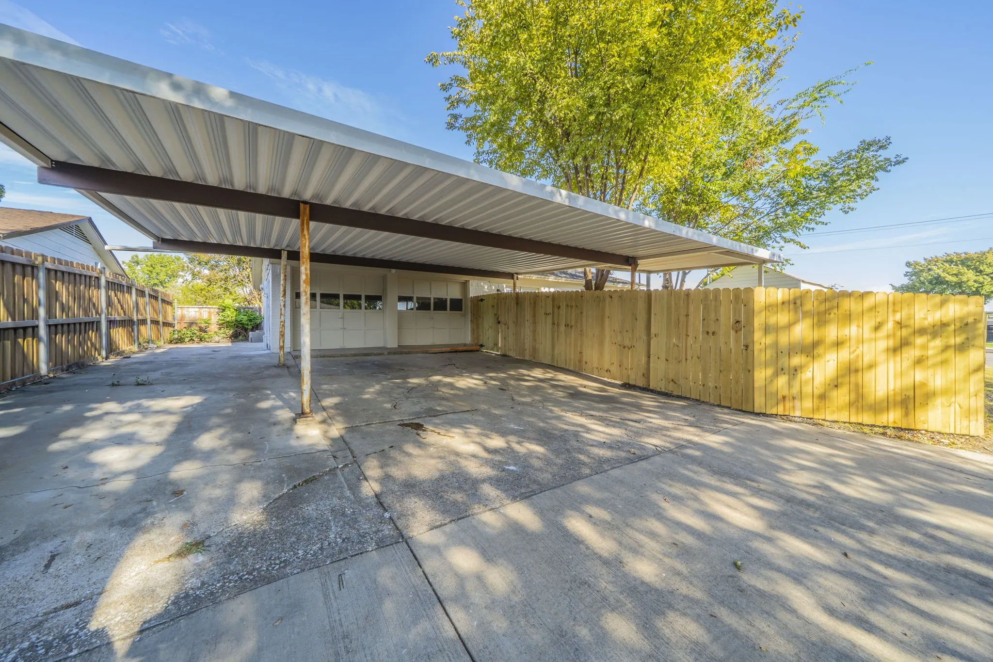 Carport with corrugated metal roofing, concrete driveway, wood privacy fencing, and an attached garage with panel doors