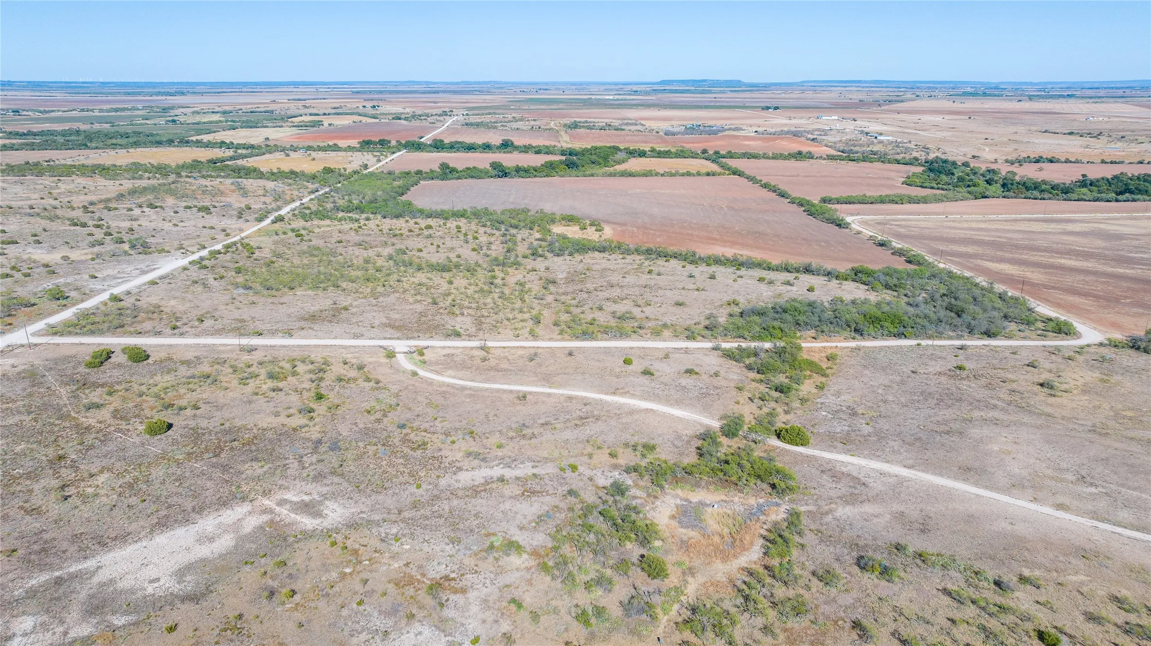 Expansive land featuring natural vegetation, visible roadways, and distant cultivated fields under a clear sky