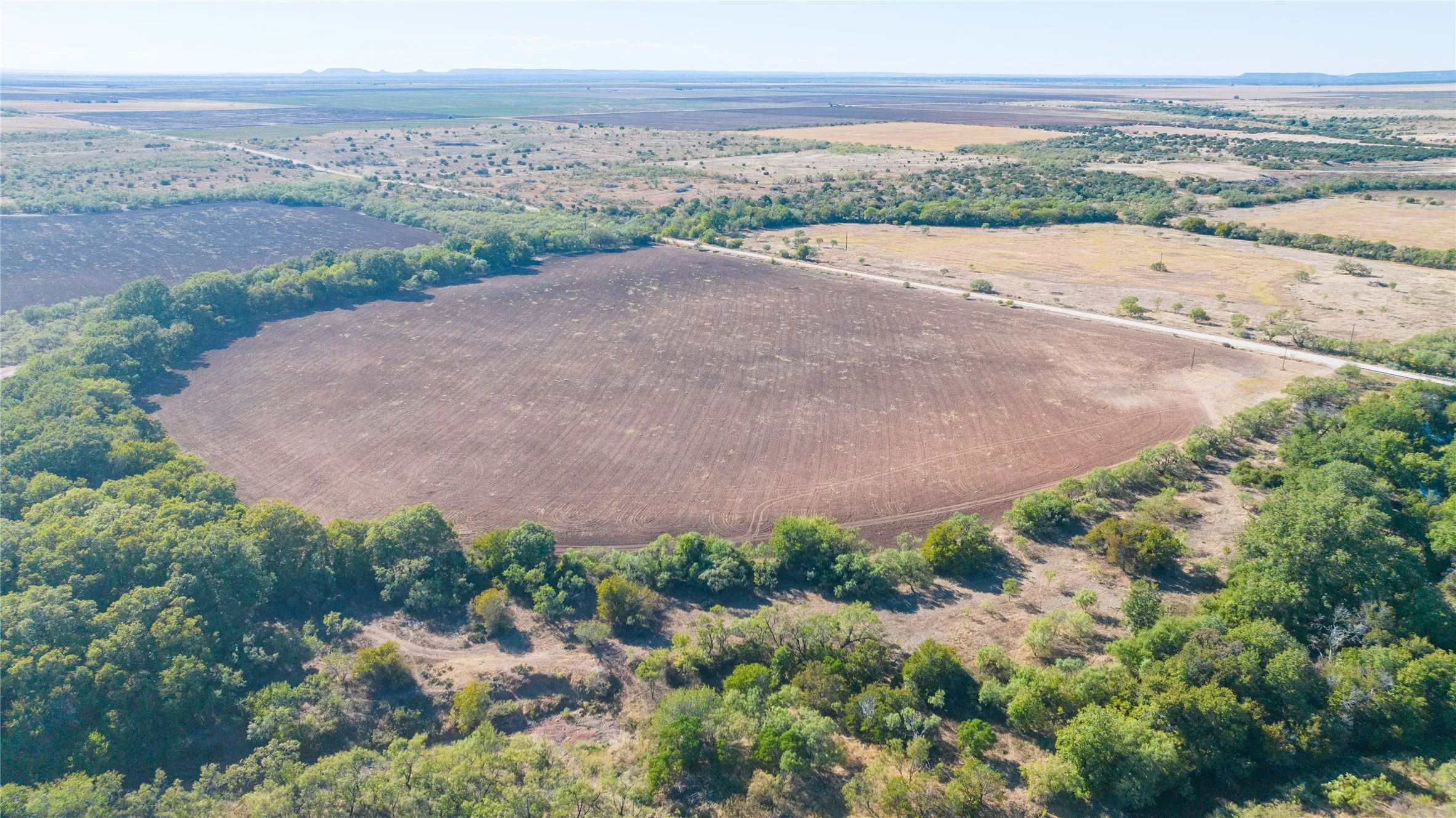 Expansive cultivated land surrounded by mature trees, featuring a clear sky and distant undeveloped terrain
