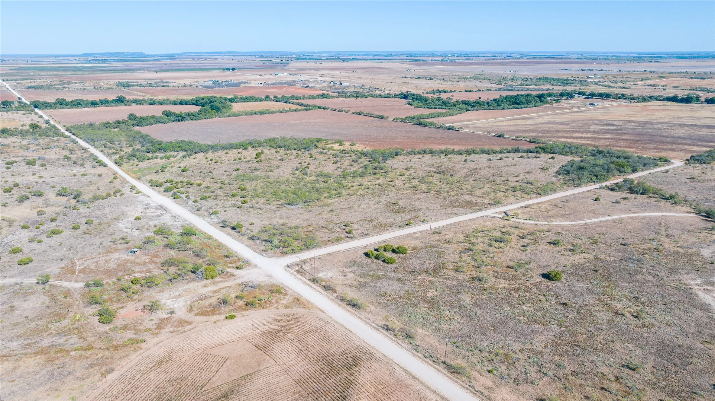 Expansive rural landscape featuring a dirt road intersection, scattered brush, and distant agricultural fields