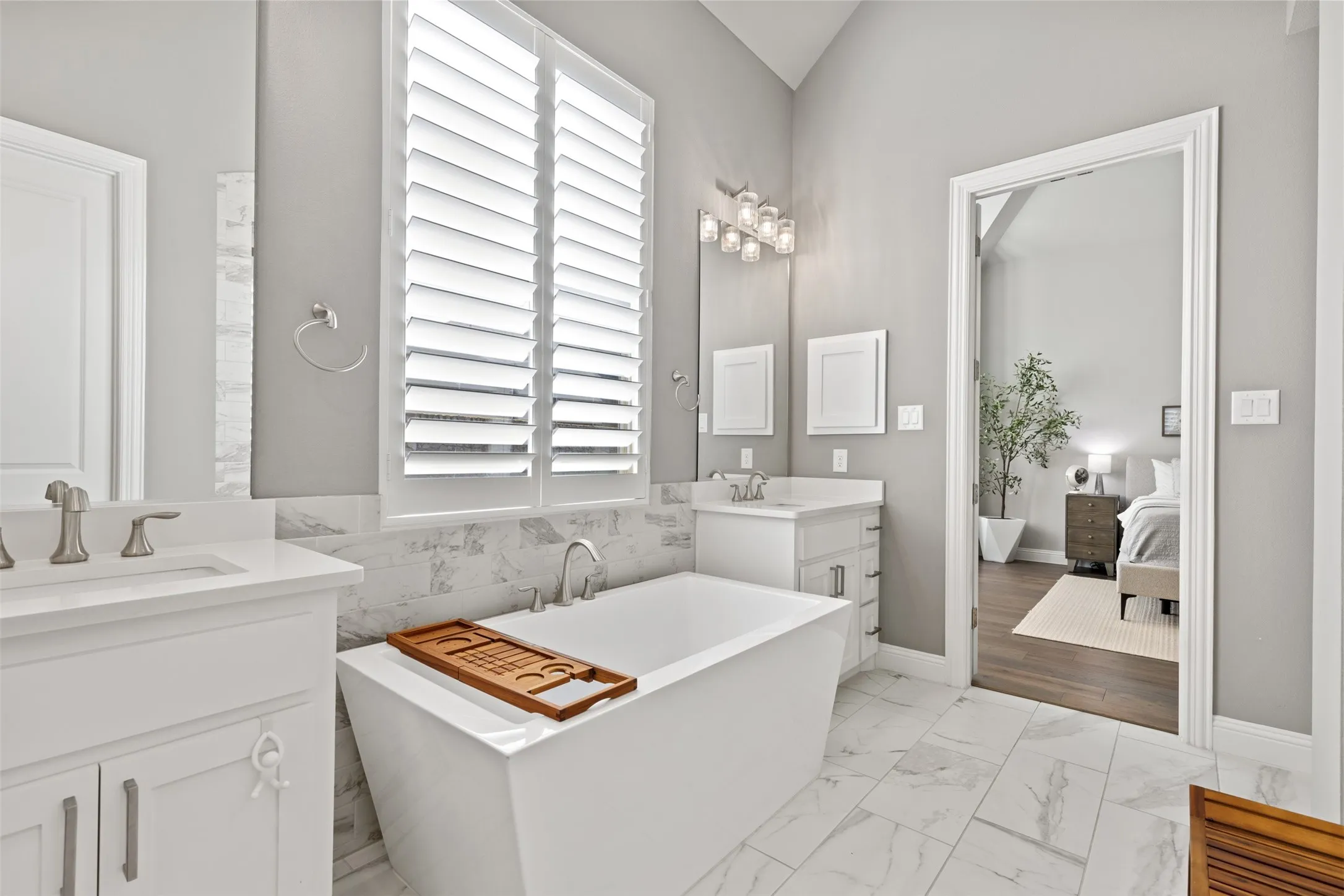 Freestanding soaking tub, white plantation shutters, dual vanities with white cabinetry, wall-mounted mirror, and tile flooring
