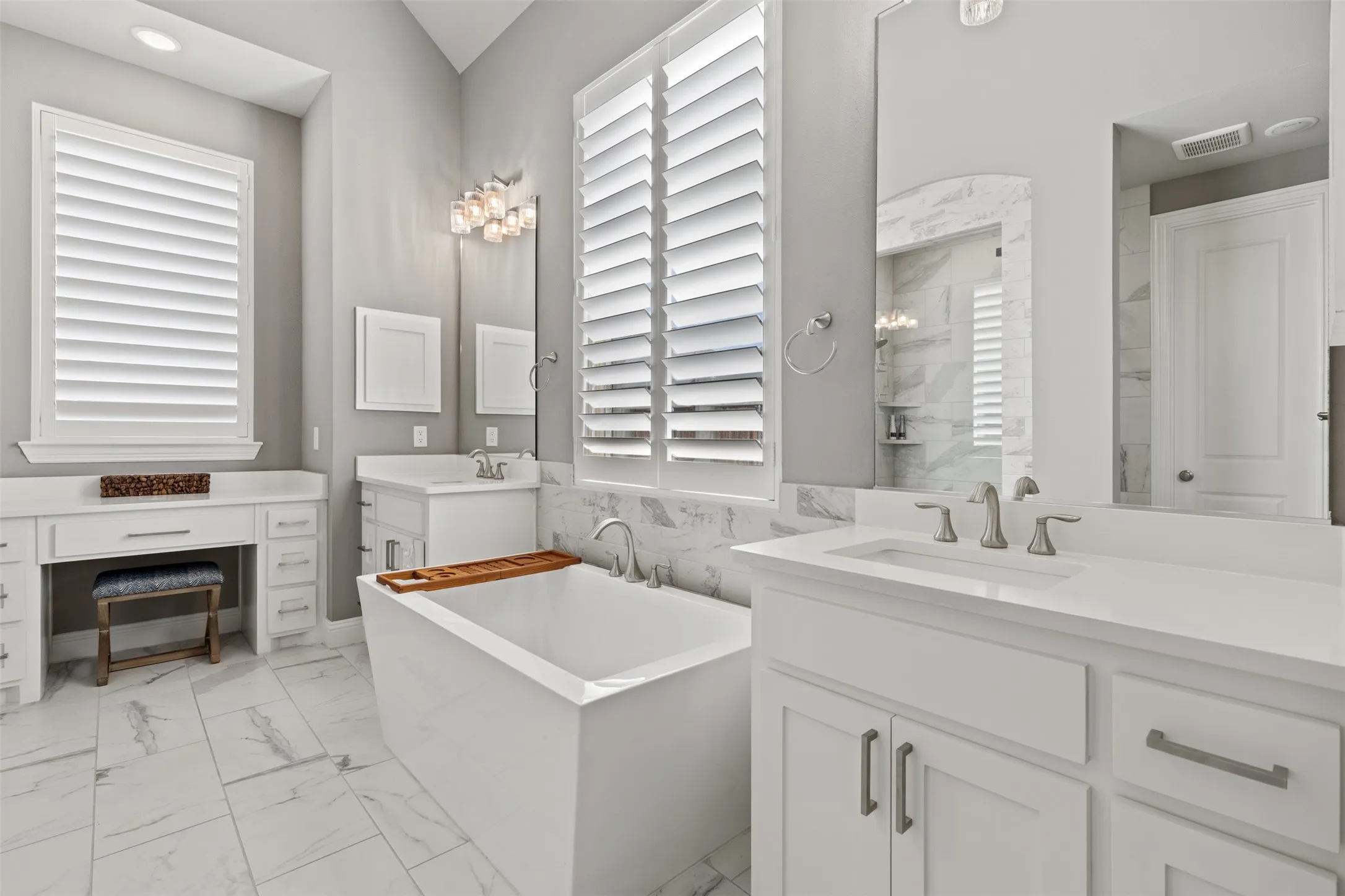 Freestanding soaking tub, dual vanity with undermount sinks, white cabinetry with brushed nickel hardware, expansive vanity counter space, and plantation shutters