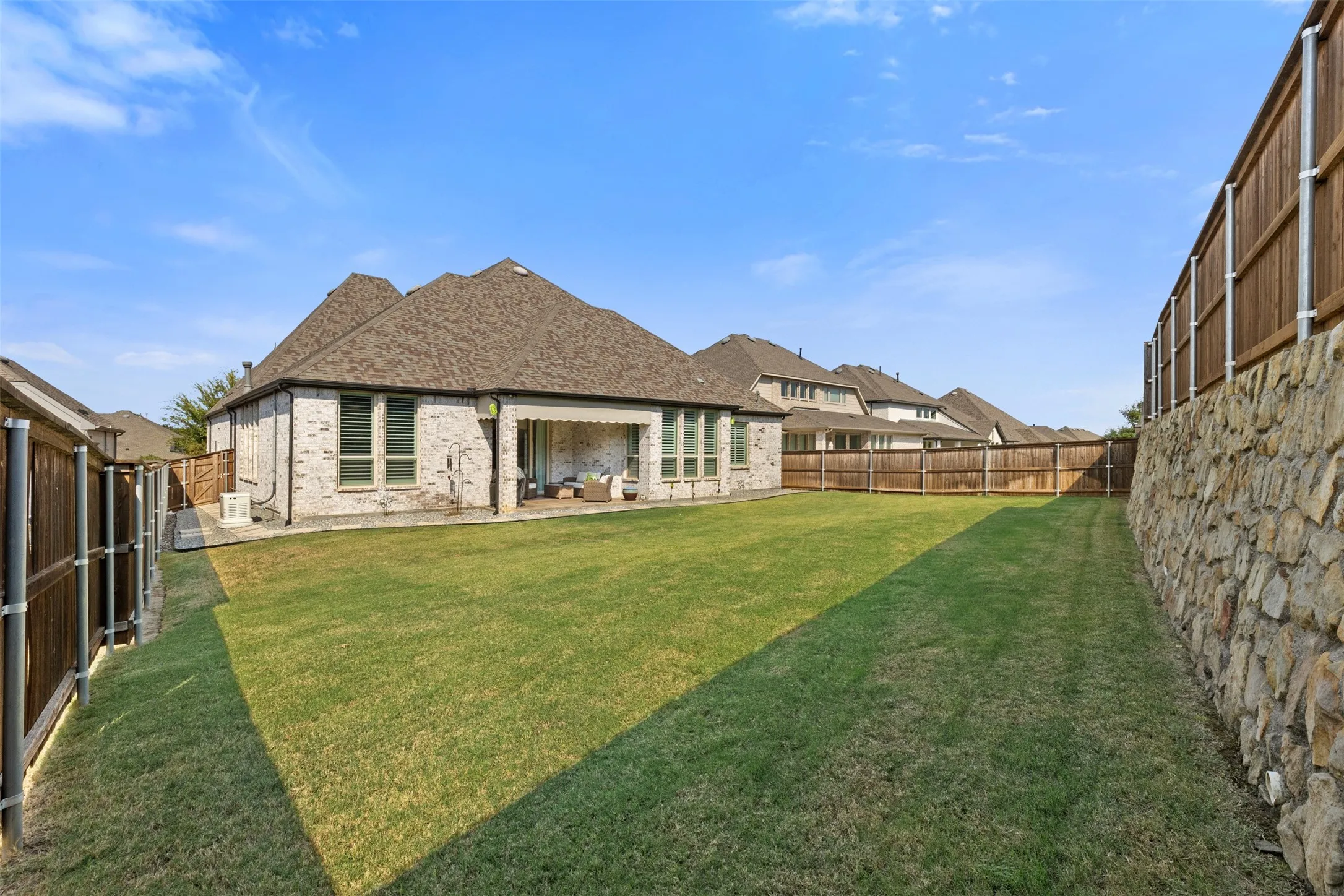 Expansive backyard featuring a manicured lawn, a stone retaining wall, and a covered patio with recessed lighting