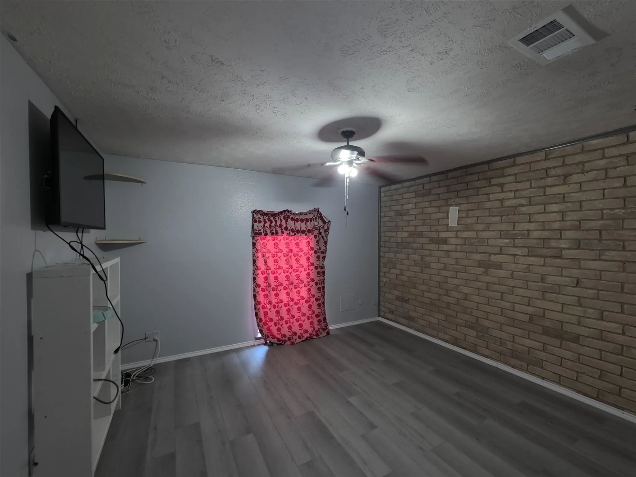 Wood-finish flooring, accent brick wall, overhead ceiling fan with lighting, and light gray wall paint