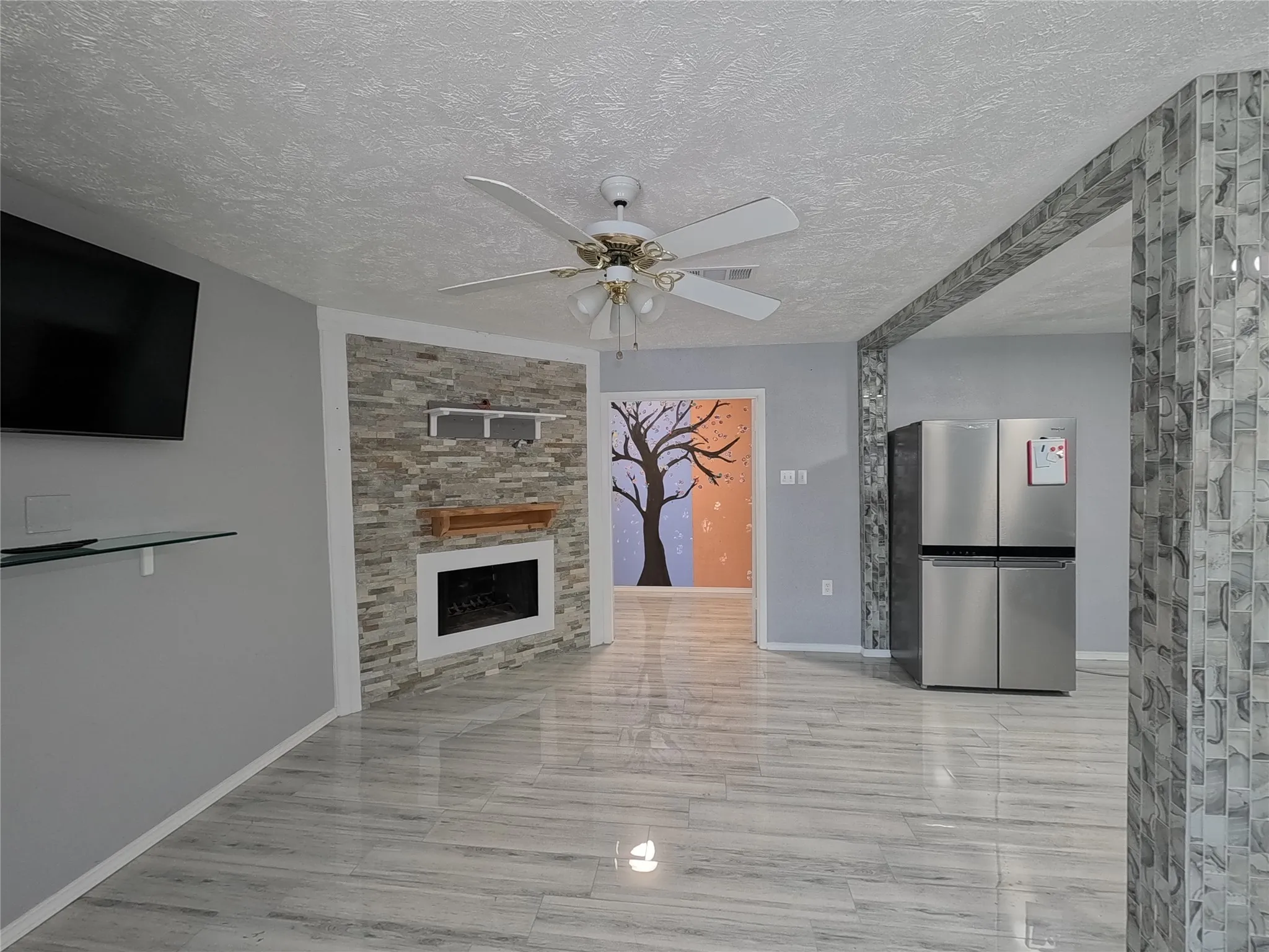 Recessed electric fireplace with a wood mantel, stacked stone accent wall, ceiling fan, and wood-finish flooring