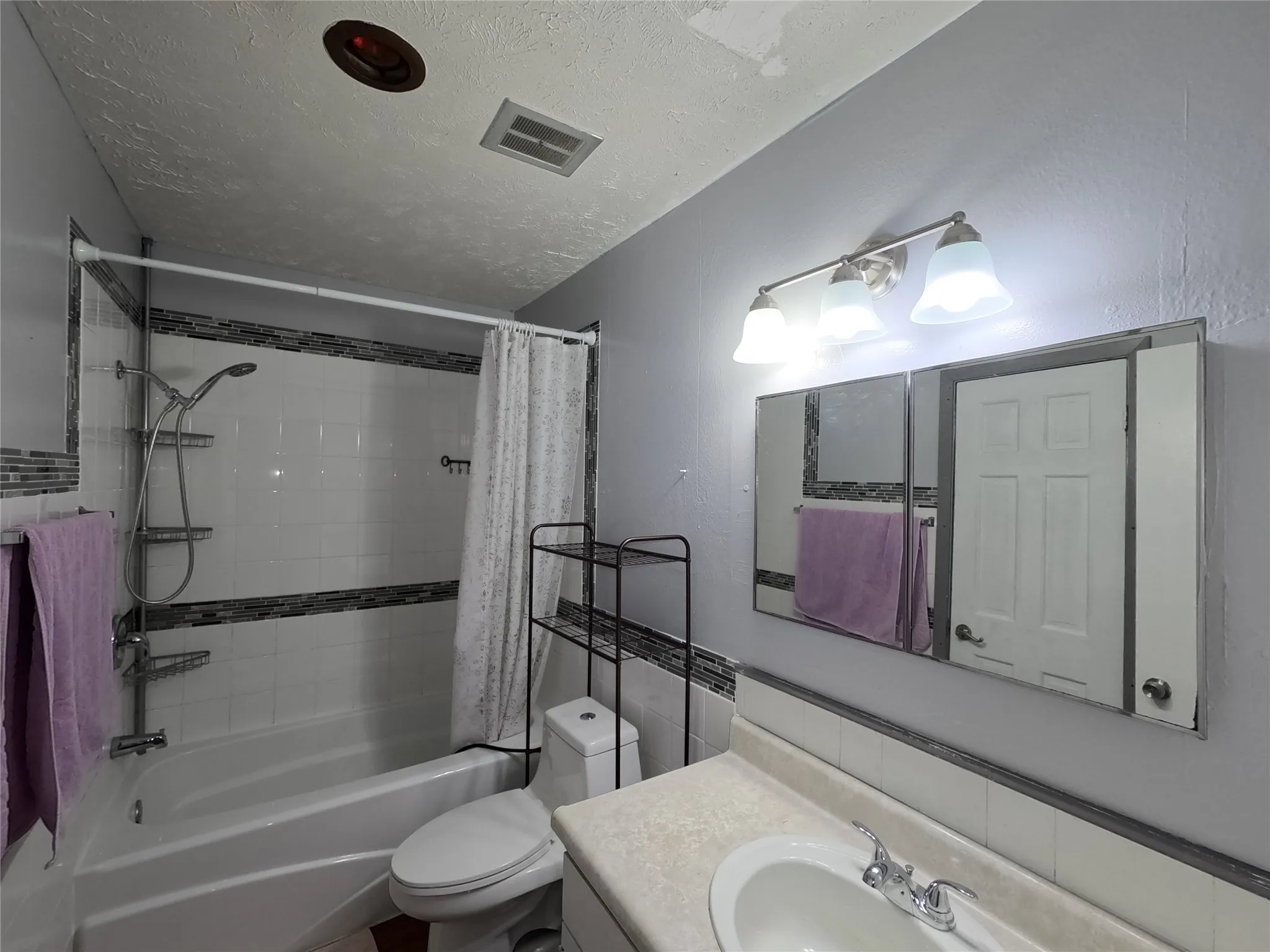 Bathtub with white tile surround and decorative accent band, chrome shower fixture, vanity with integrated sink, chrome faucet, and mirrored medicine cabinet