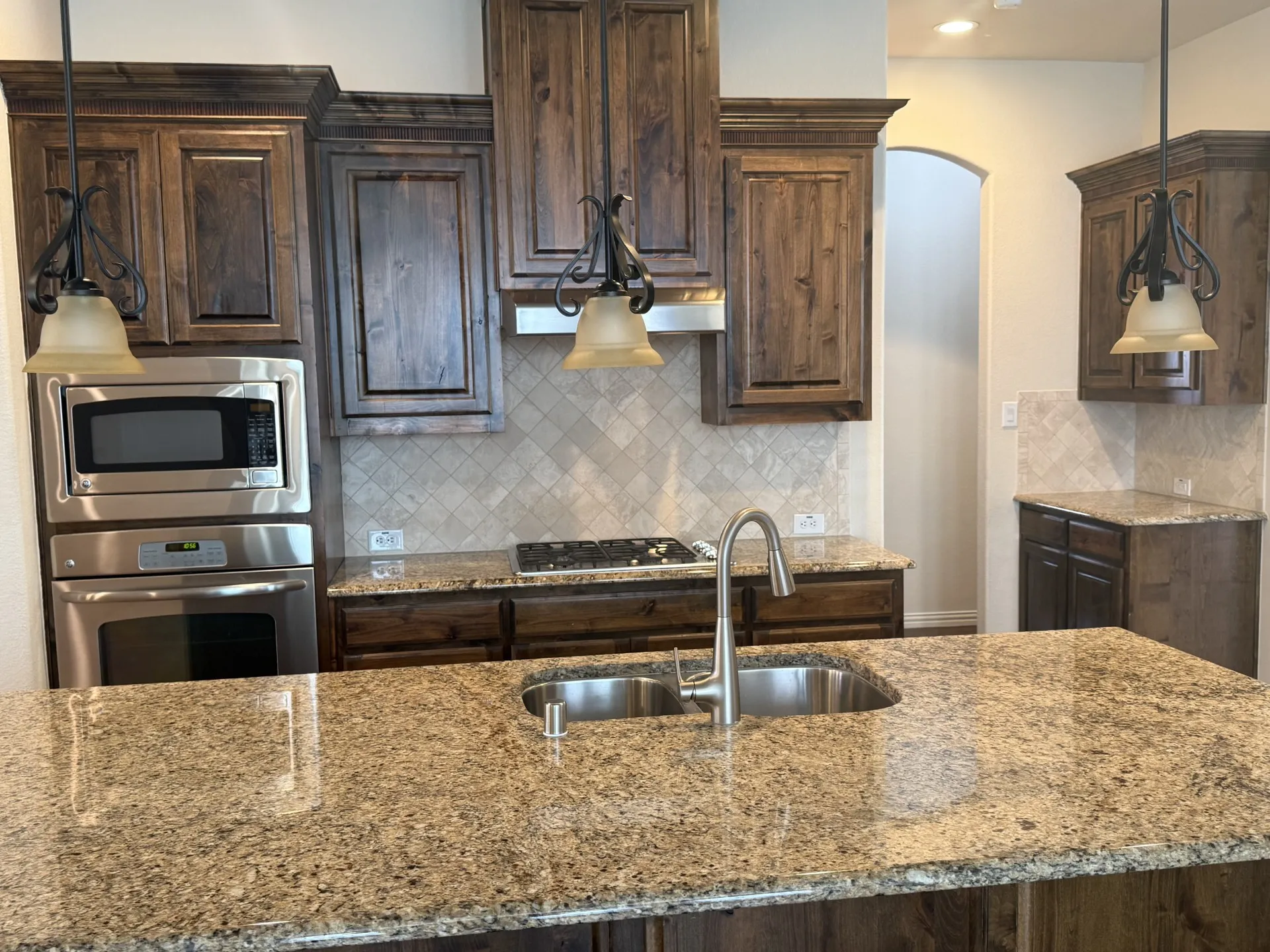 Chef's kitchen featuring custom wood cabinetry with bronze hardware, granite countertops with waterfall edge, dual stainless steel undermount sinks with gooseneck faucet, gas cooktop, and stainless steel appliances including built-in microwave and wall oven