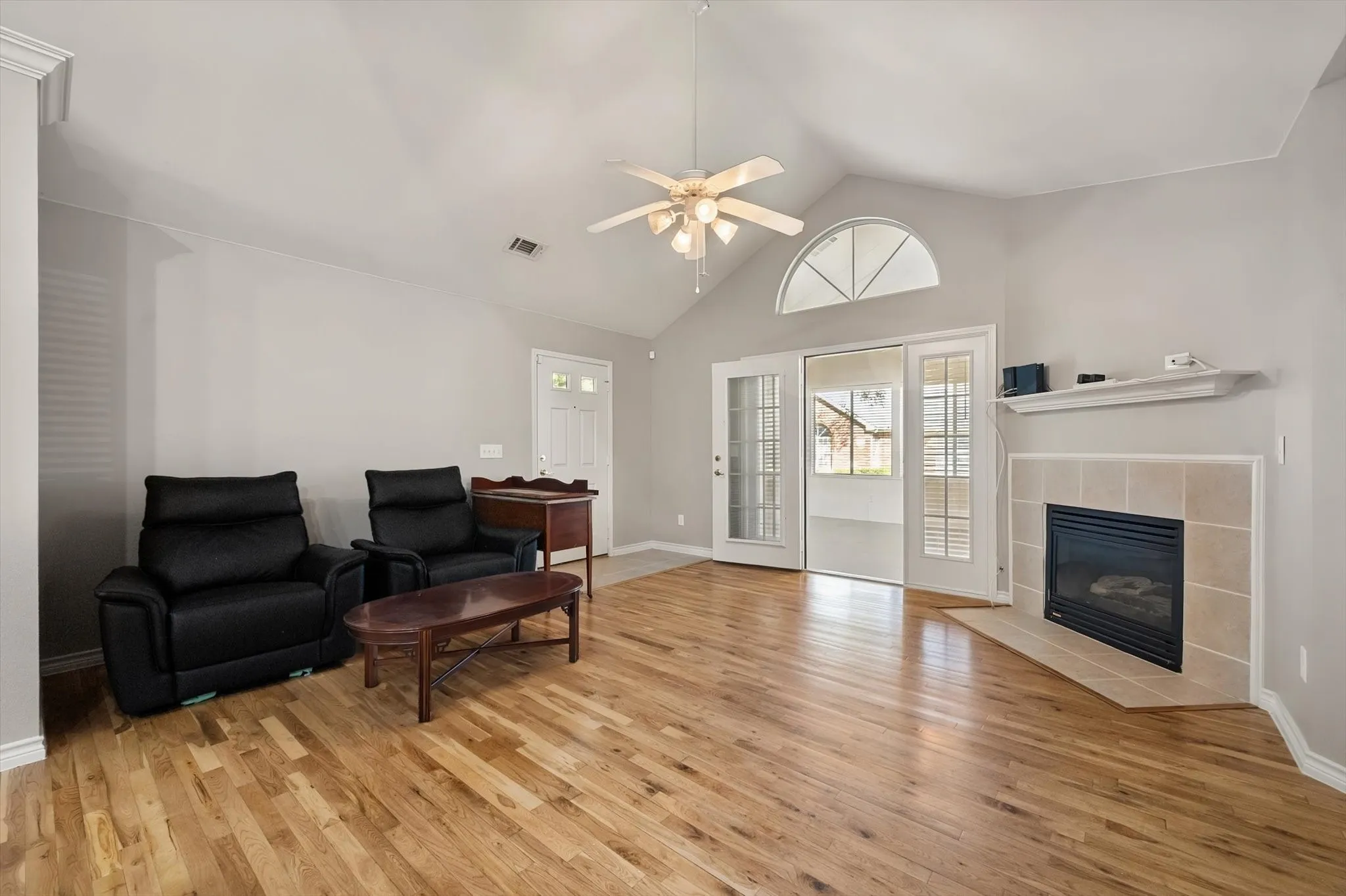 Vaulted ceiling with ceiling fan, wood-finish flooring, tiled fireplace with mantel, and glass-paneled sliding doors with transom window