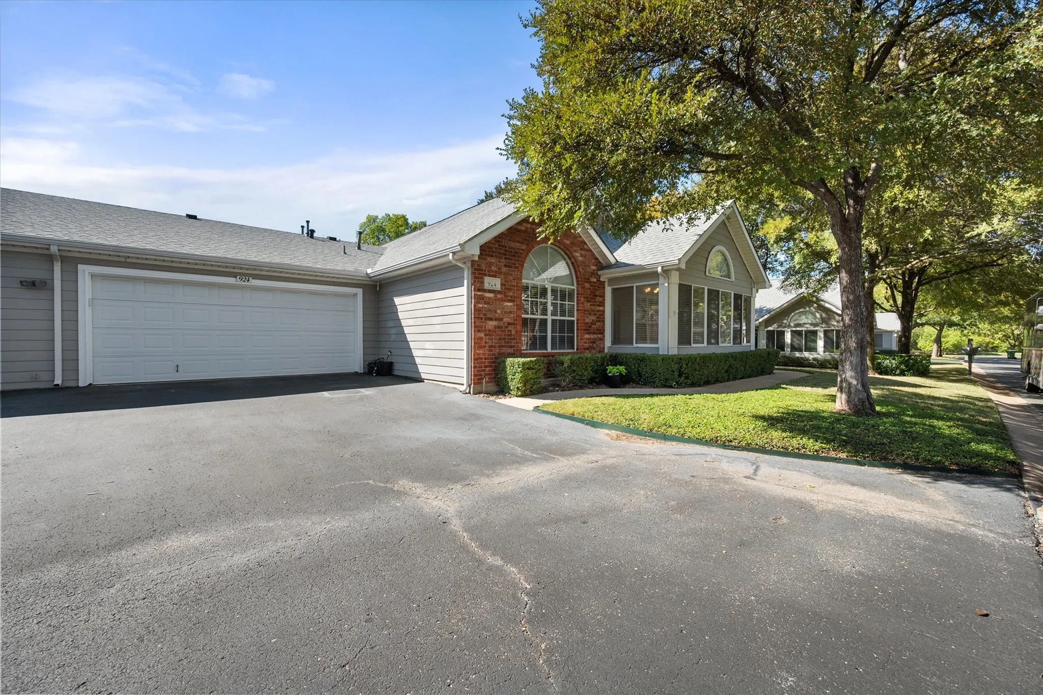 Attached two-car garage, brick facade with arched window, manicured shrubbery, and mature trees