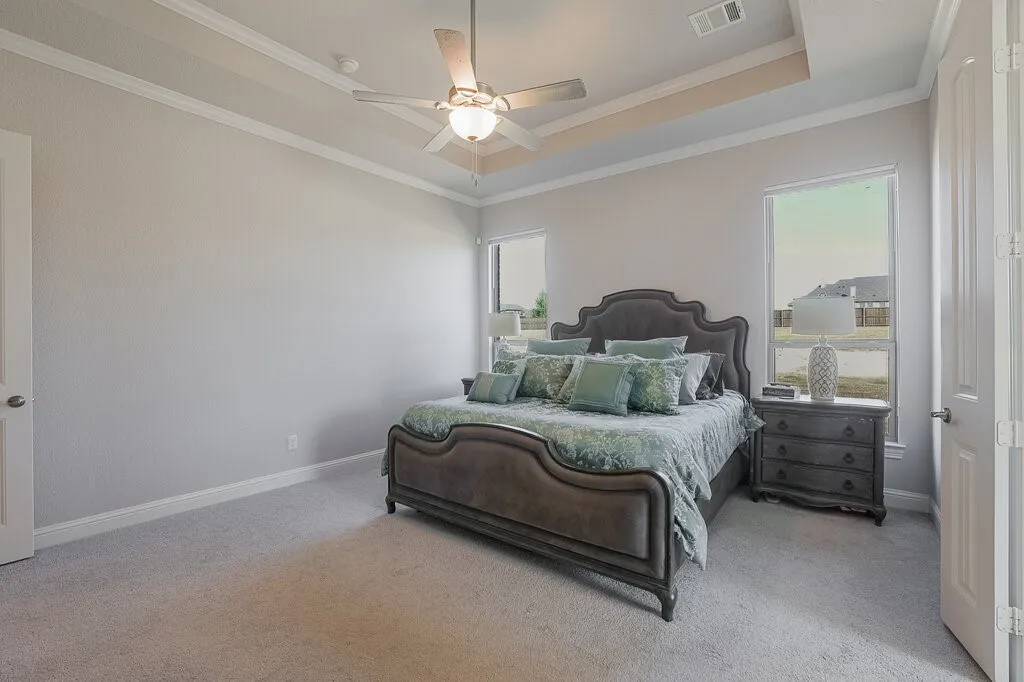 Tray ceiling with crown molding, ceiling fan with integrated lighting, two windows with white trim, and light-colored carpeting