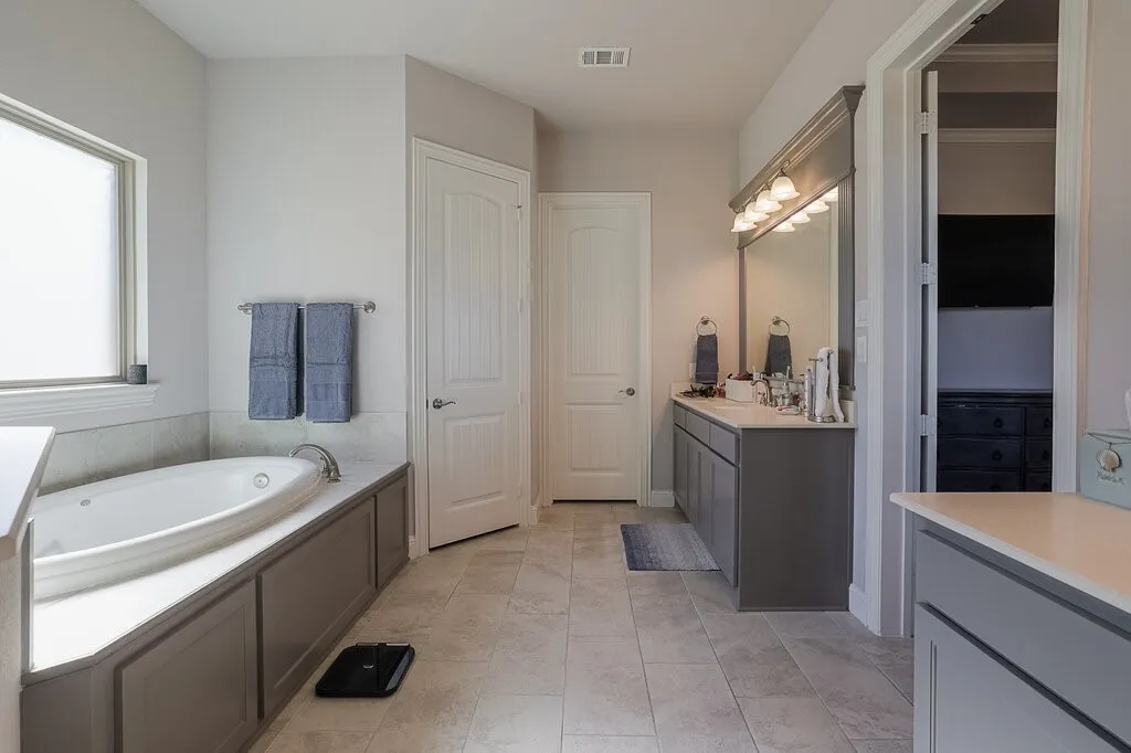 Built-in soaking tub with surround, a window with roller shade, and a vanity with dual sinks, overhead lighting, and an expansive mirror