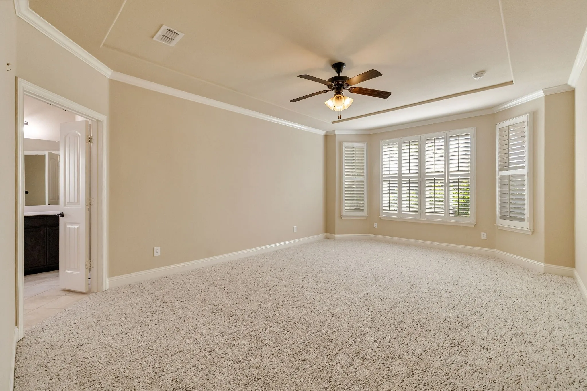 Recessed ceiling with crown molding, ceiling fan with integrated lighting, bay window with plantation shutters, and plush carpeting
