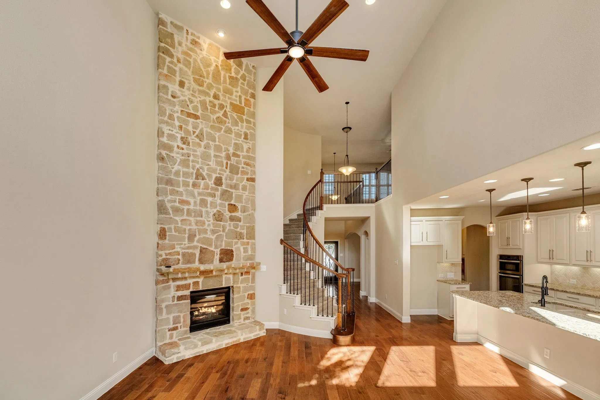 Two-story stone fireplace with a modern insert, wood-finish flooring, a spiral staircase with wood and wrought iron railings, an open-concept kitchen with white cabinetry, and recessed lighting
