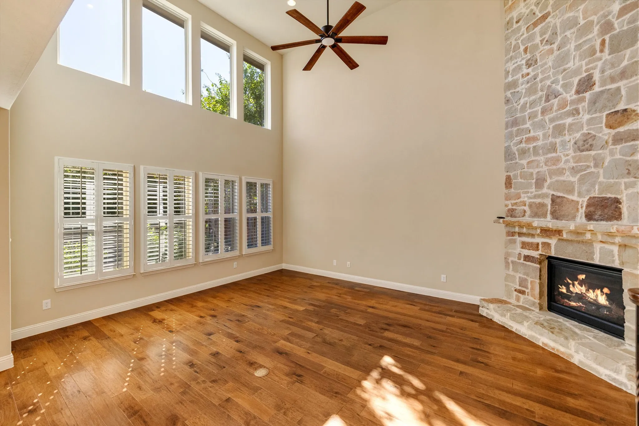 Living room featuring hardwood flooring, stone-clad fireplace with hearth, vaulted ceiling, upper transom windows, and a ceiling fan