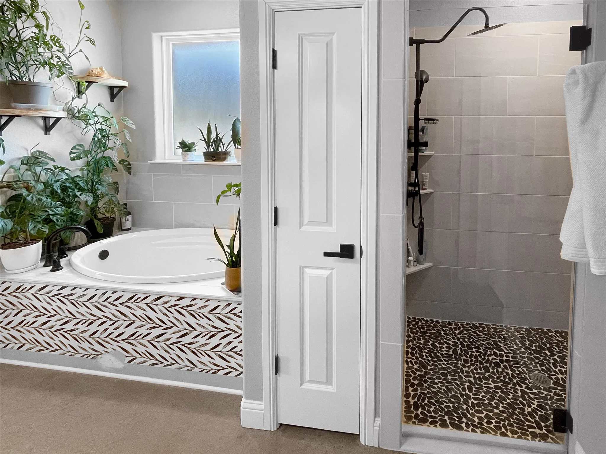Soaking tub with patterned surround, walk-in shower with pebble tile floor, matte black shower fixtures, light gray wall tile, and a frosted window