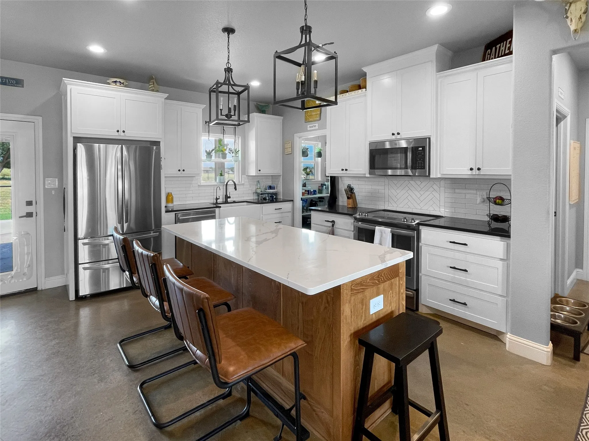 Bright kitchen featuring an expansive center island with a light-toned countertop, white cabinetry with dark hardware, a herringbone pattern tile backsplash, stainless steel appliances, and concrete flooring