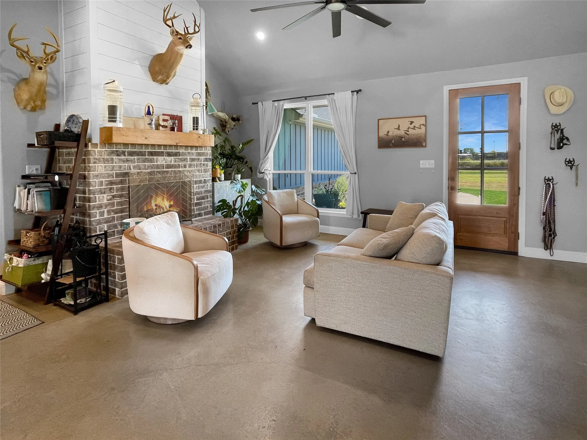 Vaulted ceiling with a ceiling fan, brick fireplace with a wood mantle, shiplap accent wall, concrete flooring, and a six-pane glass panel door